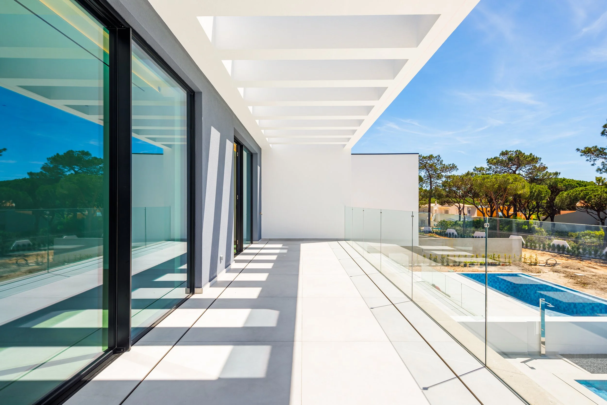 Varanda de uma casa moderna com piso de concreto branco, paredes brancas, janelas de vidro com moldura preta, e sombra projetada por uma estrutura branca acima, com vista para piscina ao ar livre e árvores verdes ao fundo.