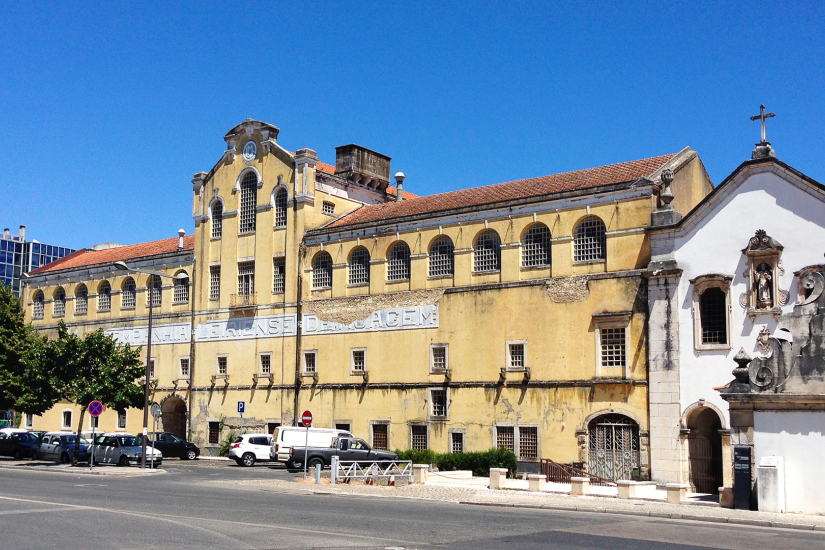 Edifício antigo com arquitetura popular, paredes amarelas e janelas pequenas, com uma fachada de igreja ao lado, sob céu azul.