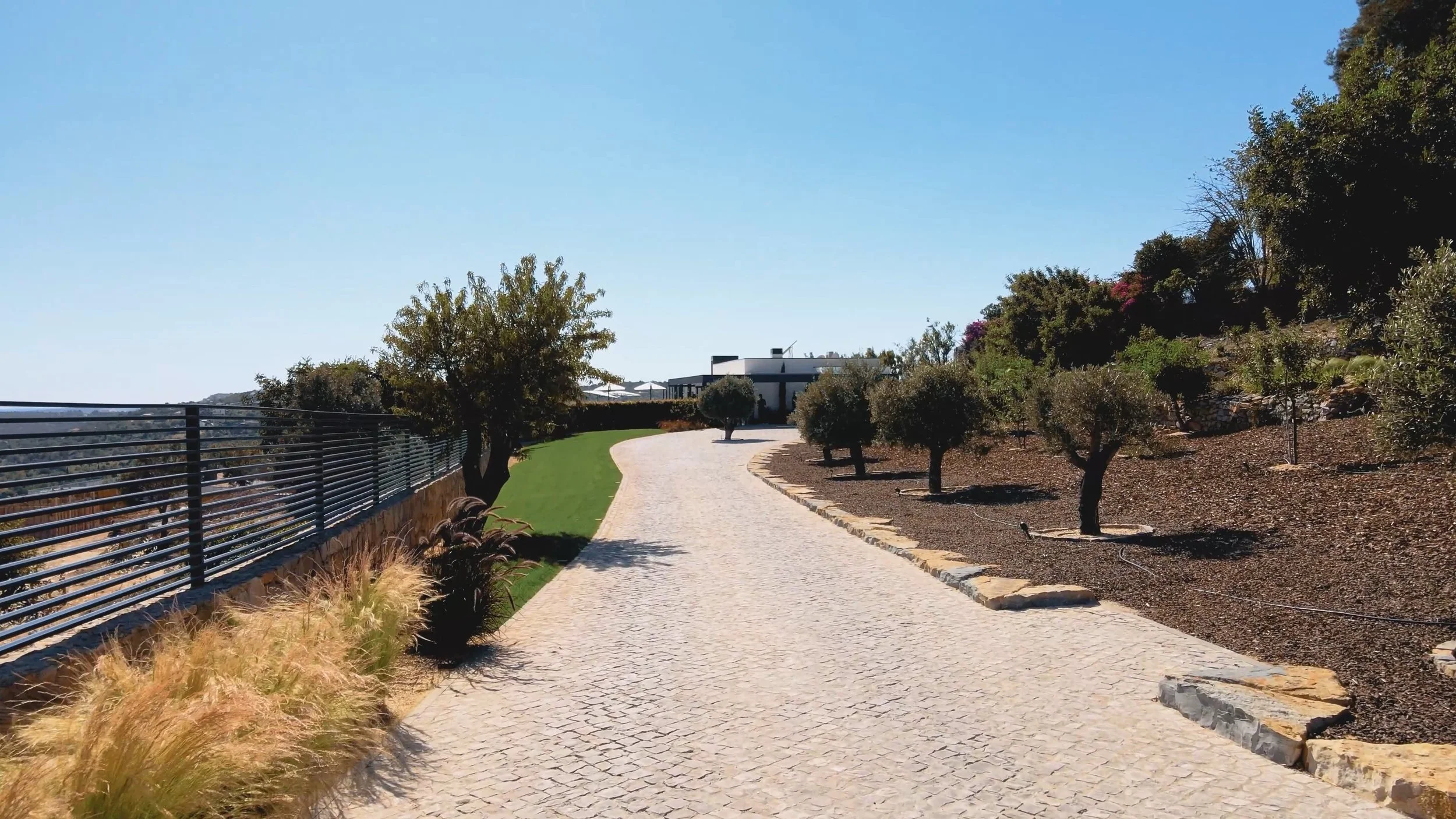 Caminho de pedra sinuoso na área externa de uma residência moderna, com árvores e arbustos ao redor, sob céu claro e ensolarado.