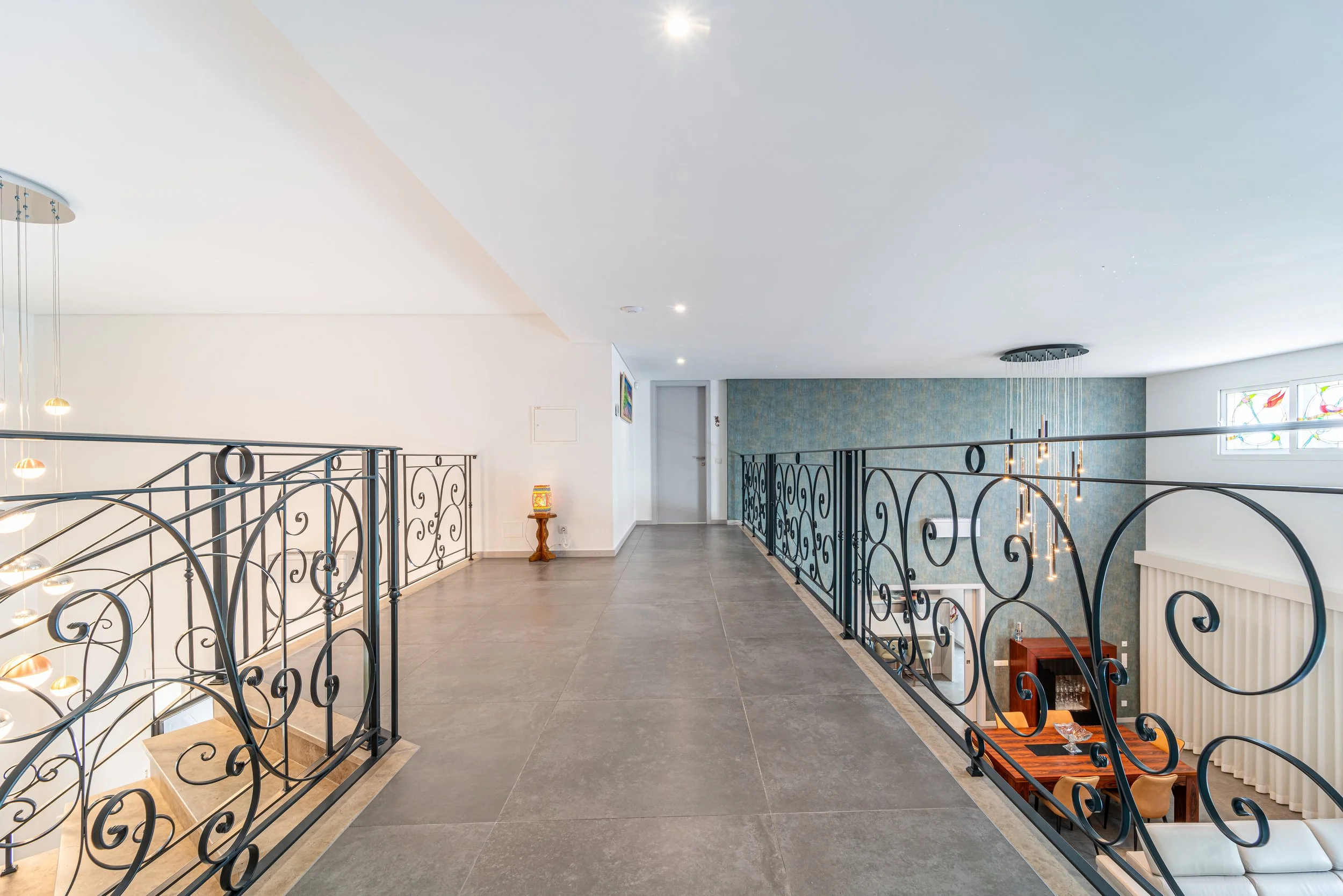 Mezanino com corrimãos de ferro com detalhes em espiral, piso de azulejo cinza, parede azul com textura e janelas com vitrais coloridos.