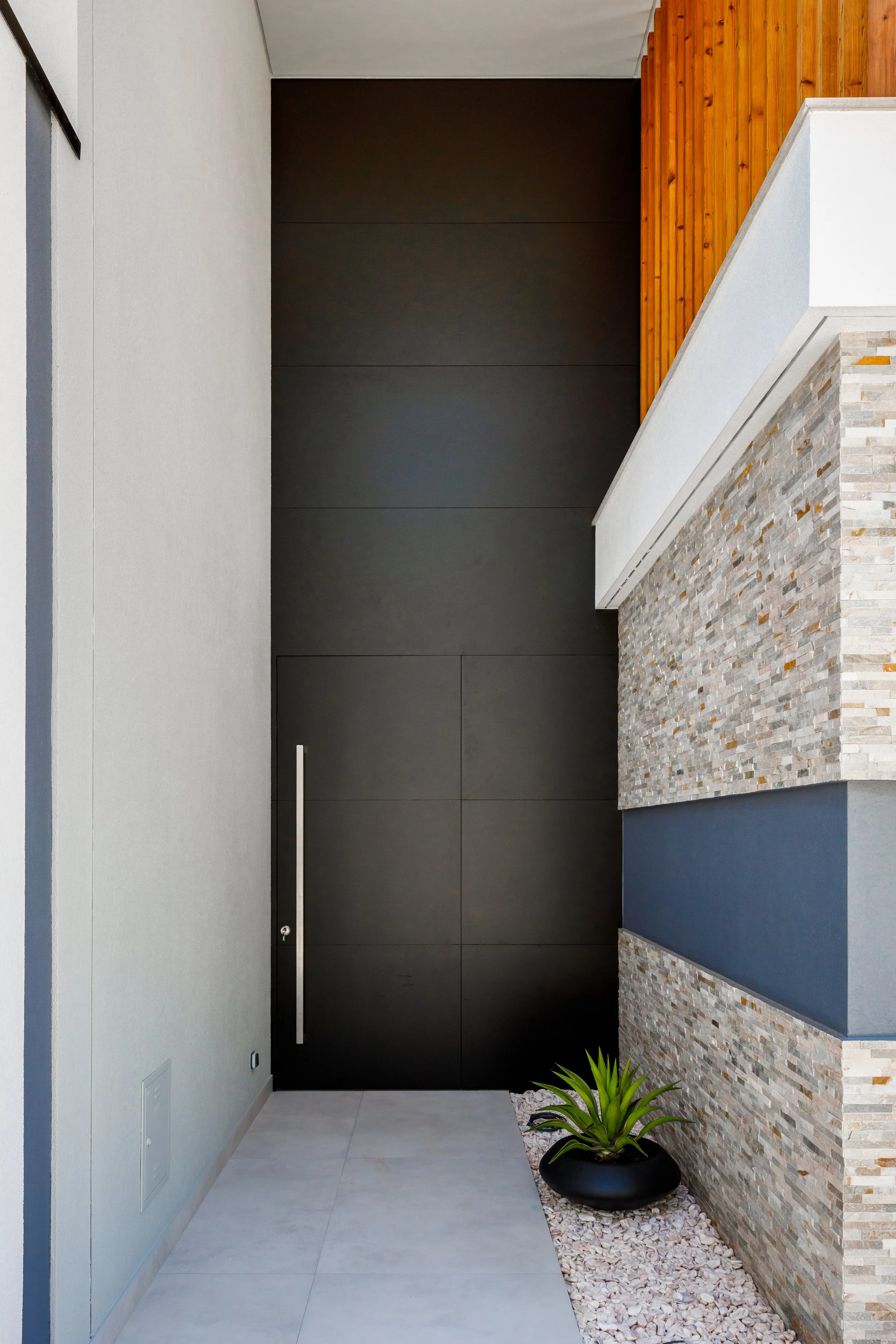 Entrada de uma casa com parede preta, parede de pedra, parede branca e uma planta em um vaso preto.