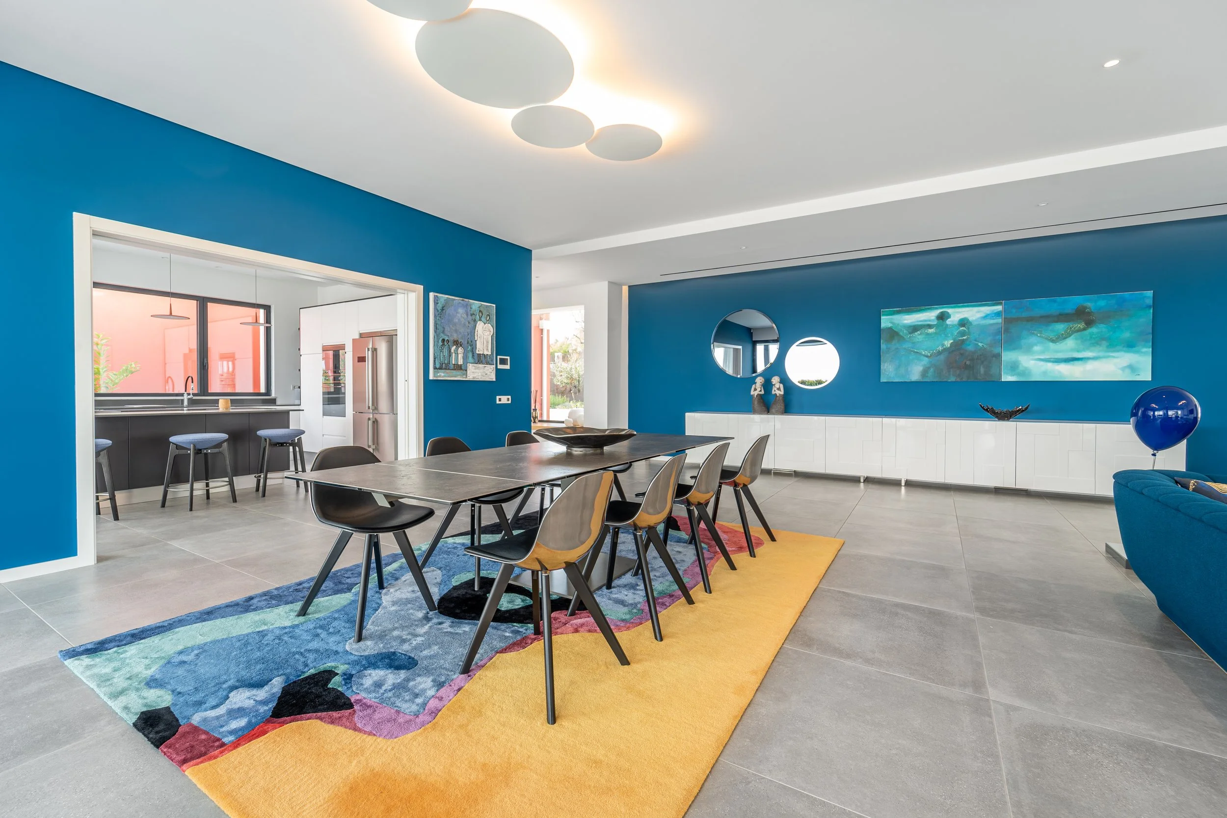 Sala de jantar moderna com paredes azuis, mesa retangular com cadeiras pretas e de madeira, tapete colorido sob a mesa, articulos decorativos, um sofá azul com balão azul, e uma parede com quadros, espelhos e esculturas.