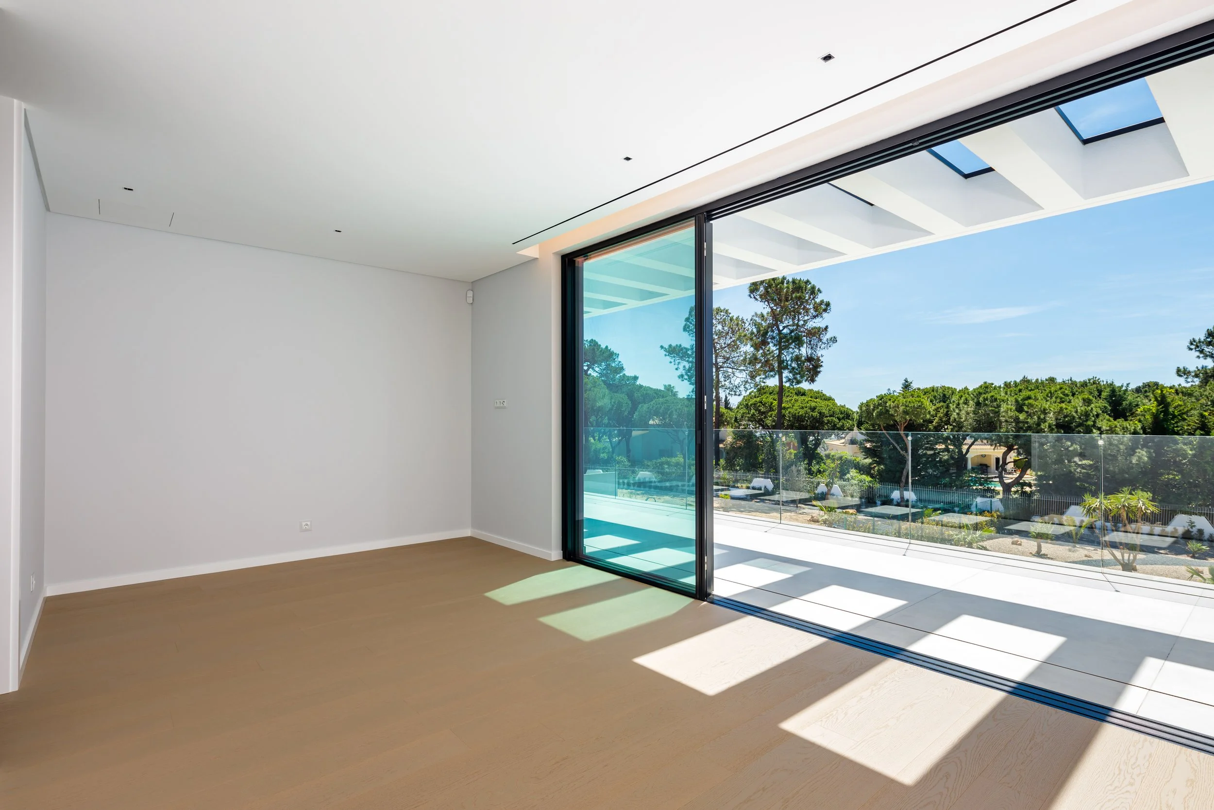 Sala vazia com piso de madeira clara, paredes brancas e porta de vidro que leva a uma varanda com vista para árvores verdes.