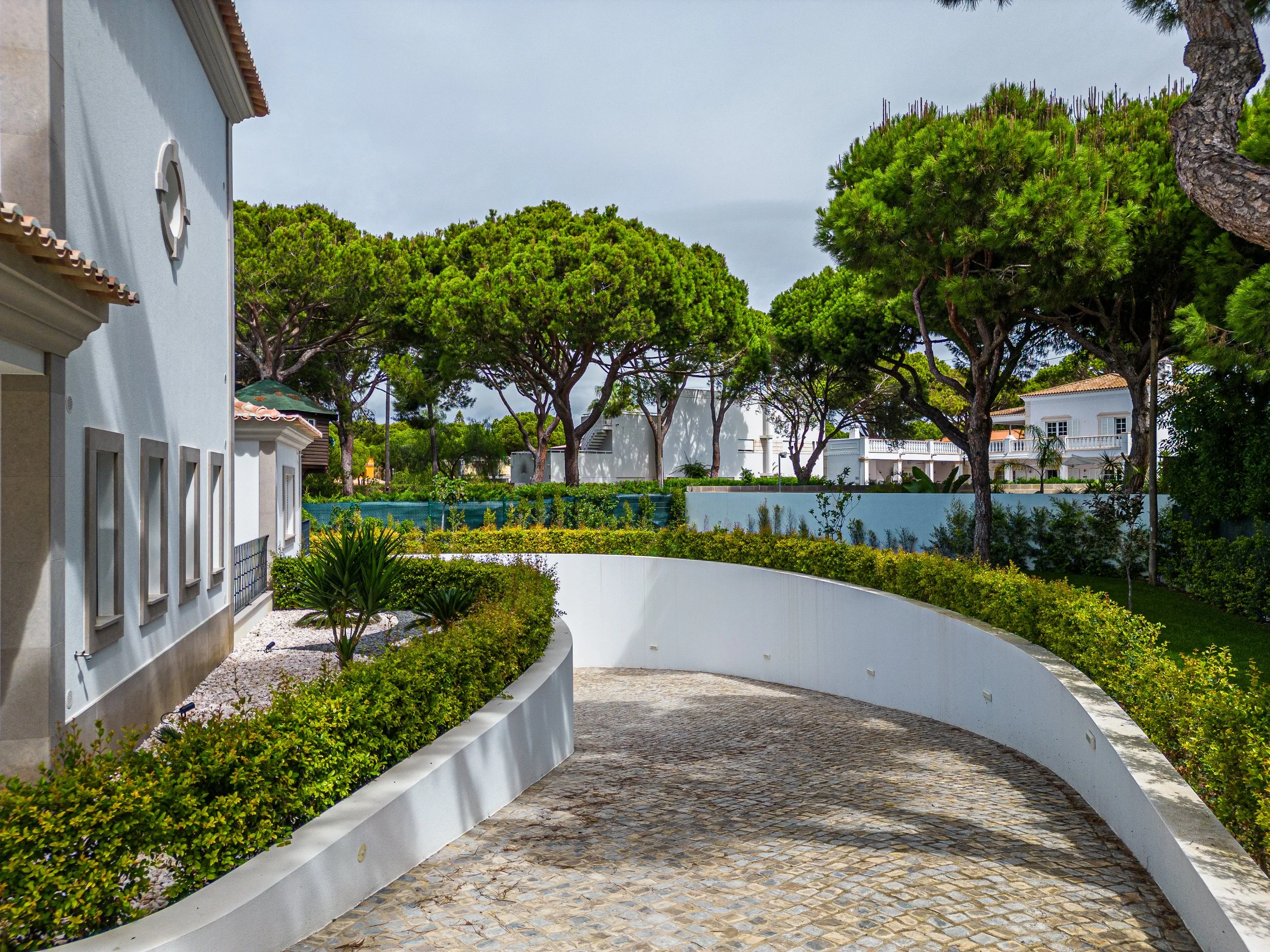 Caminho de calçamento de pedras curvas ao lado de jardim com arbustos verdes e árvores altas ao fundo. Edifícios brancos ao redor.