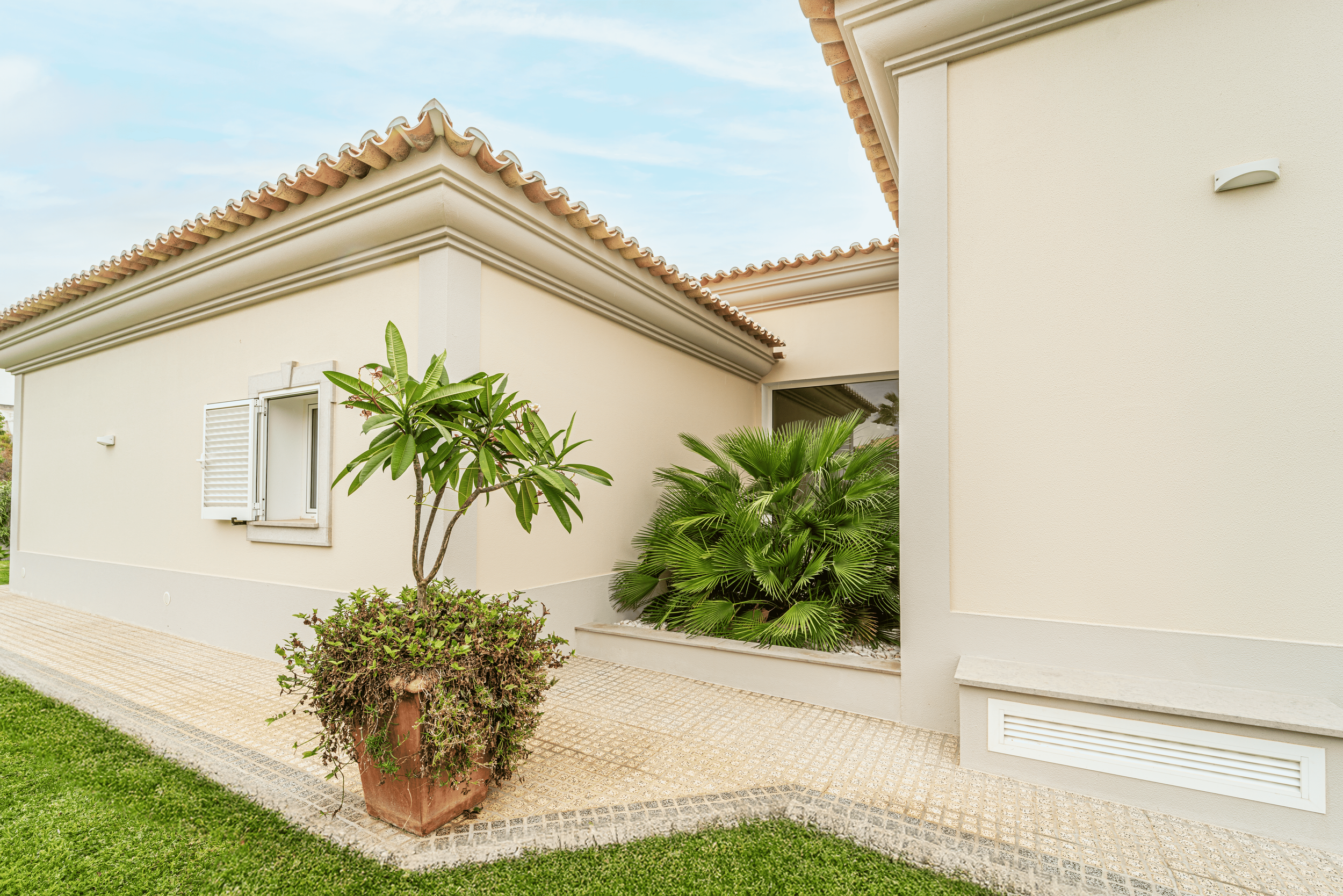 Fachada de uma casa com paredes claras, telhado de telhas e plantas no jardim, incluindo uma árvore em um vaso e folhagens verdes
