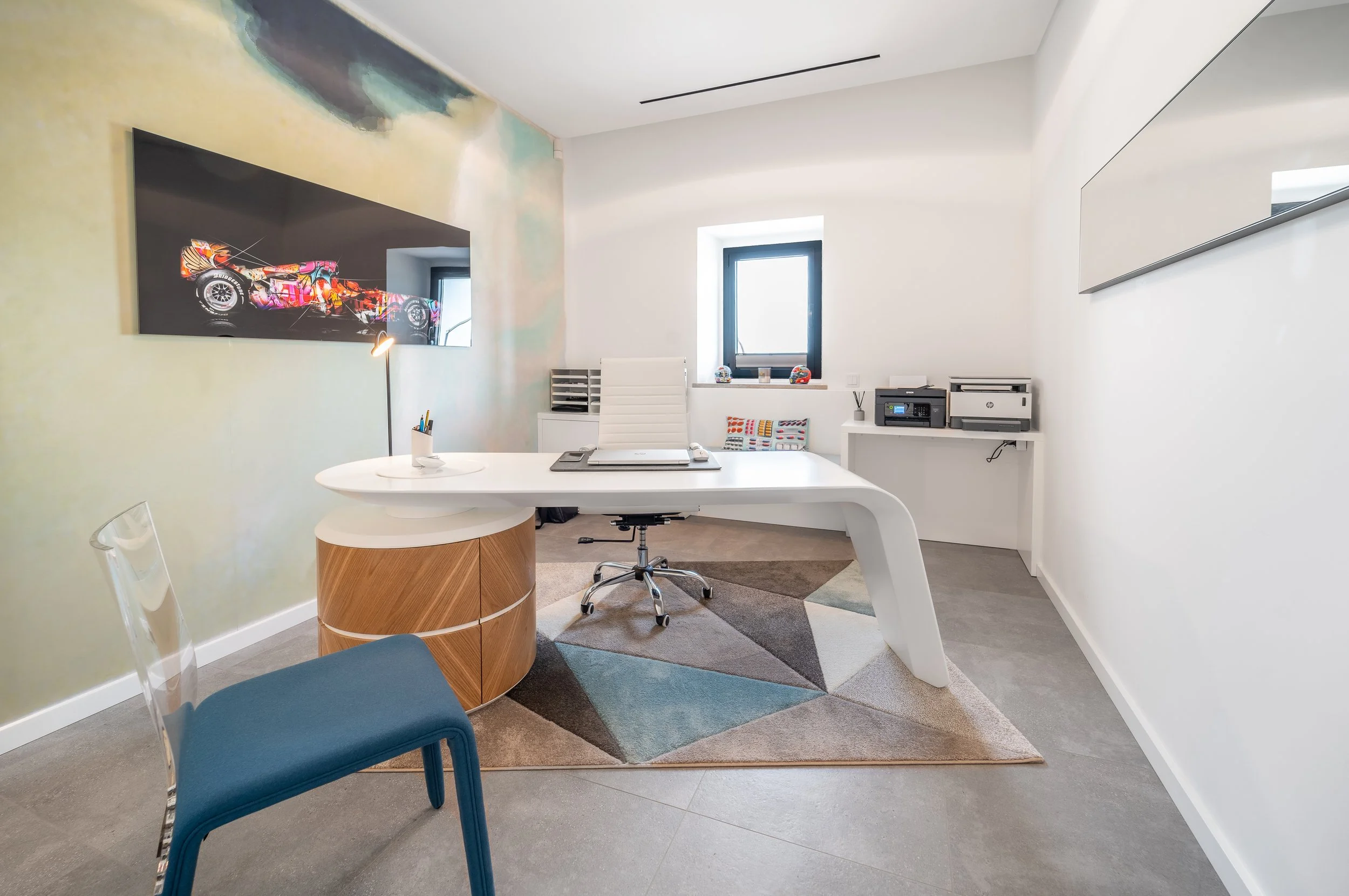 Sala de escritório moderna com mesa branca, cadeira branca, poltrona azul, quadro na parede, janela com vista para o exterior, e armário branco com impressoras.