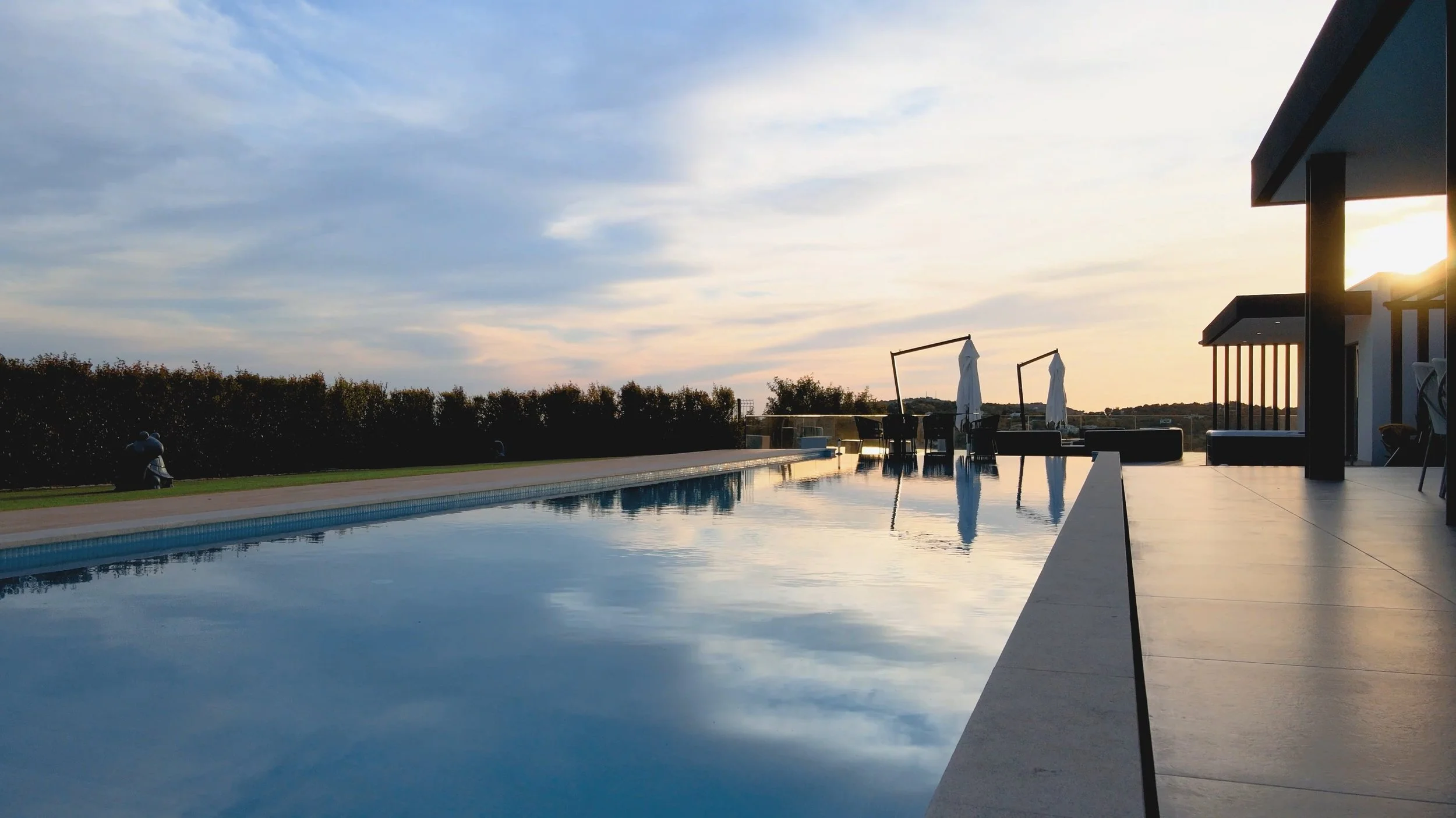 Piscina ao pôr do sol com espreguiçadeiras e guarda-sóis ao fundo, perto de uma área de lazer ao ar livre.