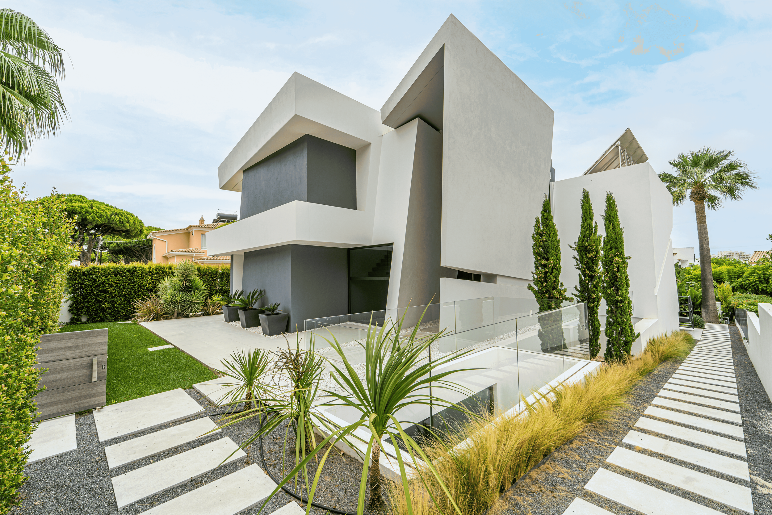 Edifício moderno branco com design geométrico, cercado por jardim bem cuidado, plantas e árvores, caminho de pedras, cerca de vidro e céu claro.