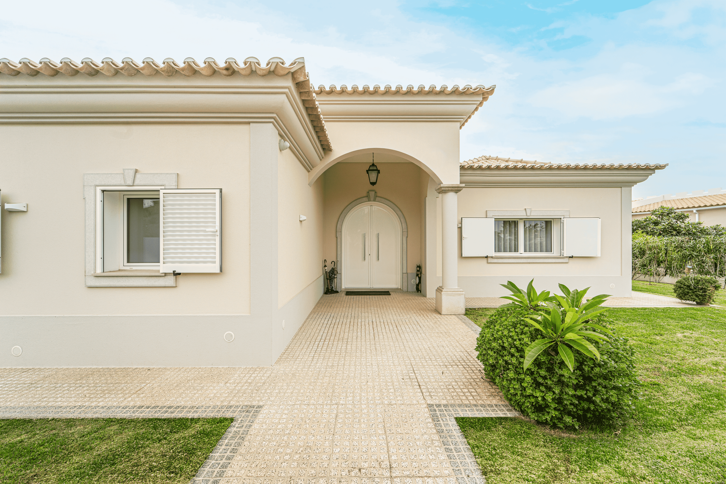 Fachada de casa com entrada principal, jardim bem cuidado, janela com persiana branca, porta de entrada dupla sob arco, teto de telhas aparentes e céu azul com algumas nuvens.