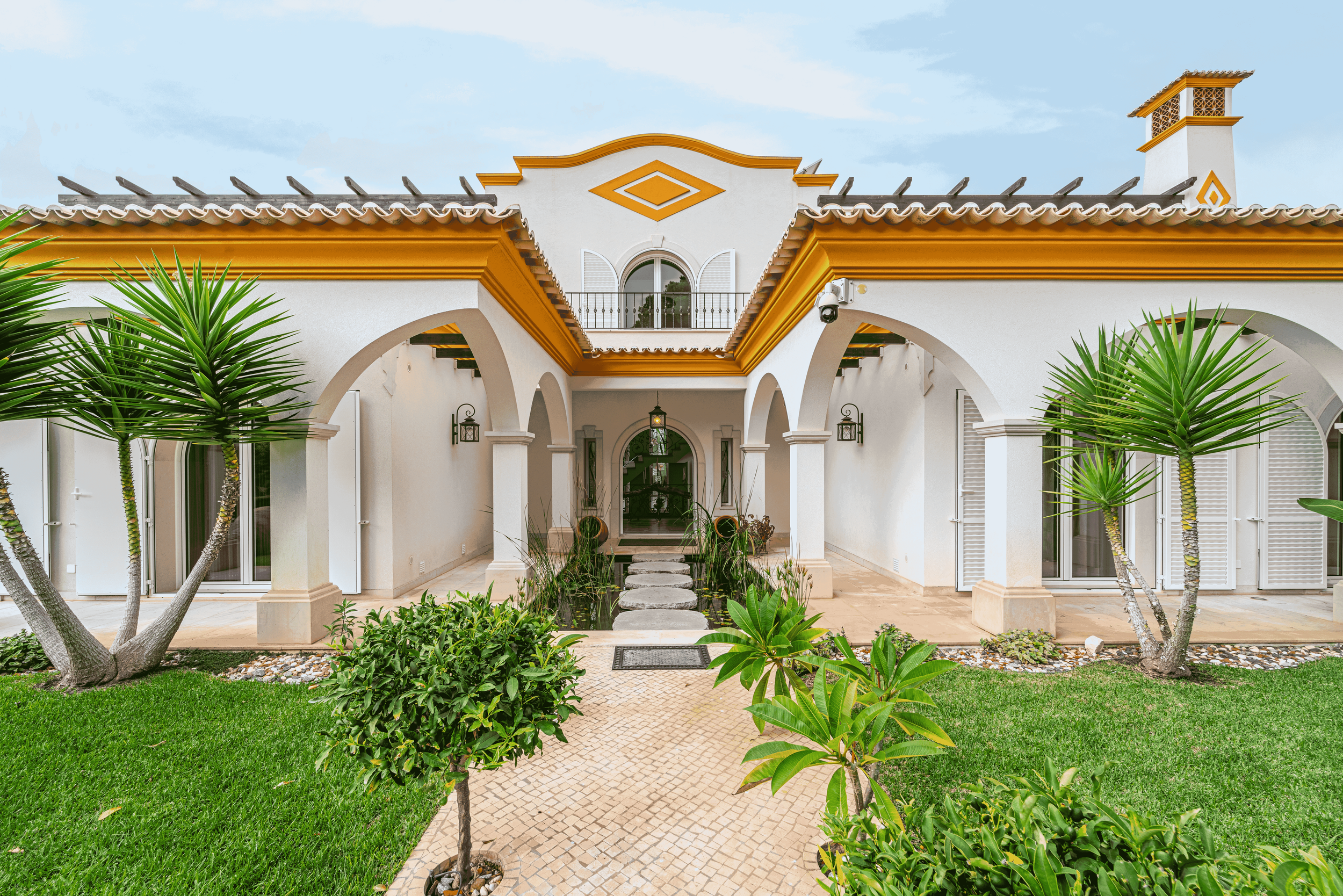 Residência com fachada branca, detalhes em amarelo, jardim bem cuidado com árvores e plantas, caminho de pedras no centro, portas de vidro e janelas com persianas, varanda com guarda-corpo de ferro, teto de telhas cerâmicas.