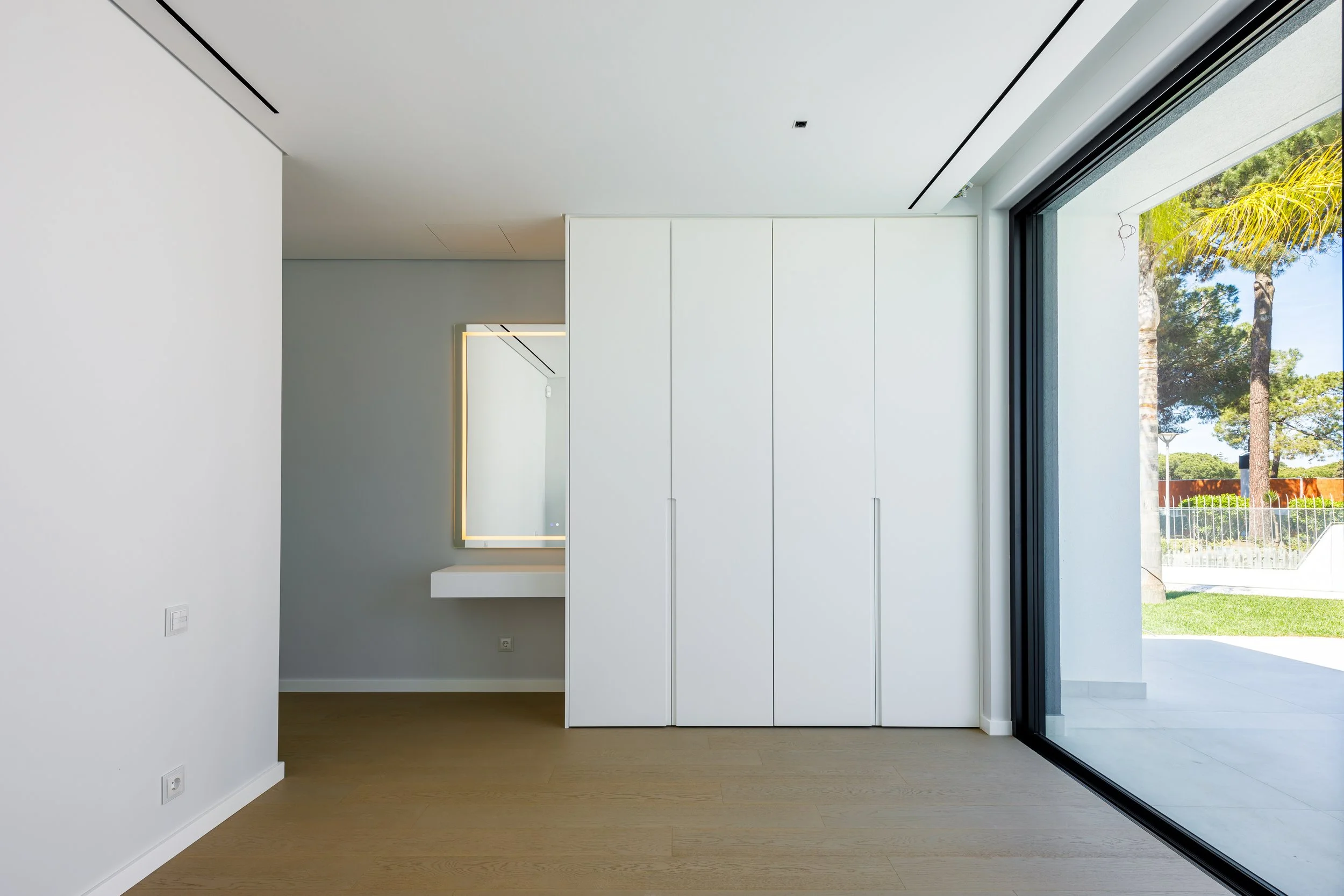 Interior de sala moderna com armário branco e espelho com iluminação ao fundo, grande janela de vidro oferece vista para jardim com árvores.