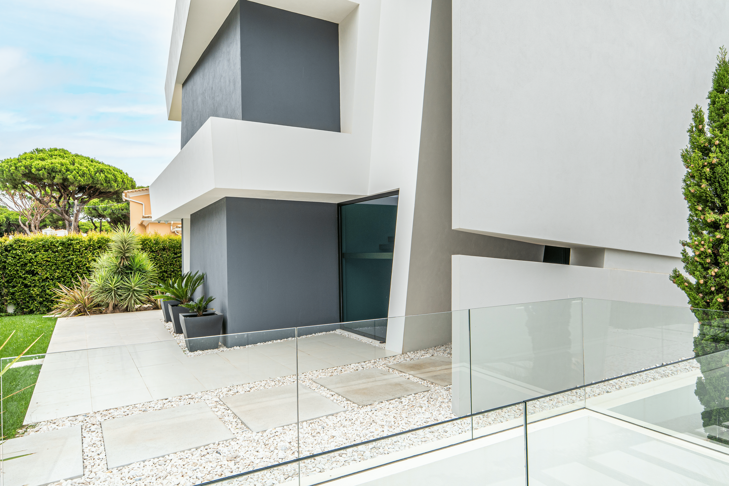 Fachada moderna de casa com paredes brancas e cinza, jardim com plantas suculentas e parede de vidro na varanda.