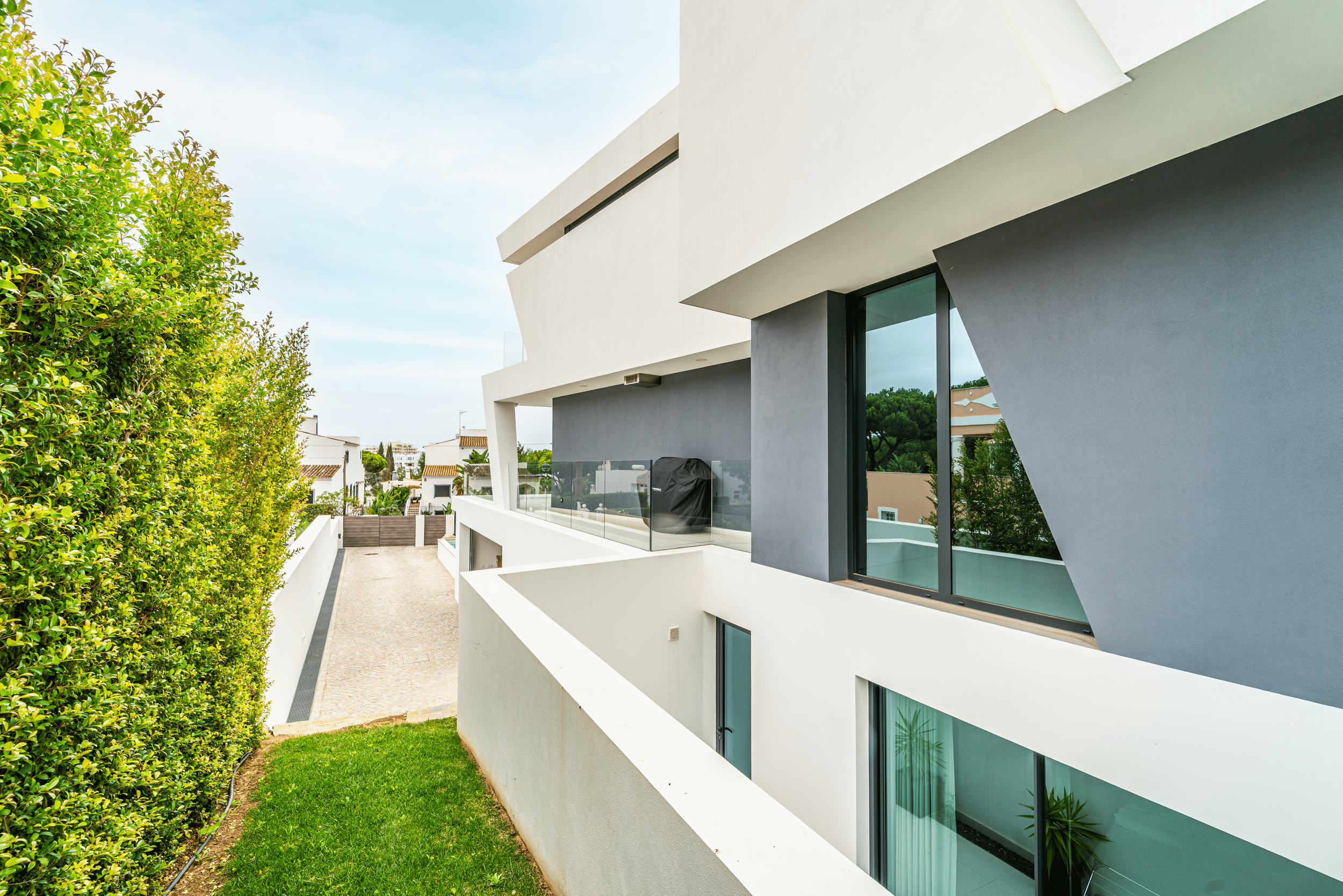 Fachada de um prédio moderno com janelas grandes de vidro, paredes brancas e cinzas, uma varanda com parede de vidro e um espaço de jardim com grama e uma sebe verde ao lado.