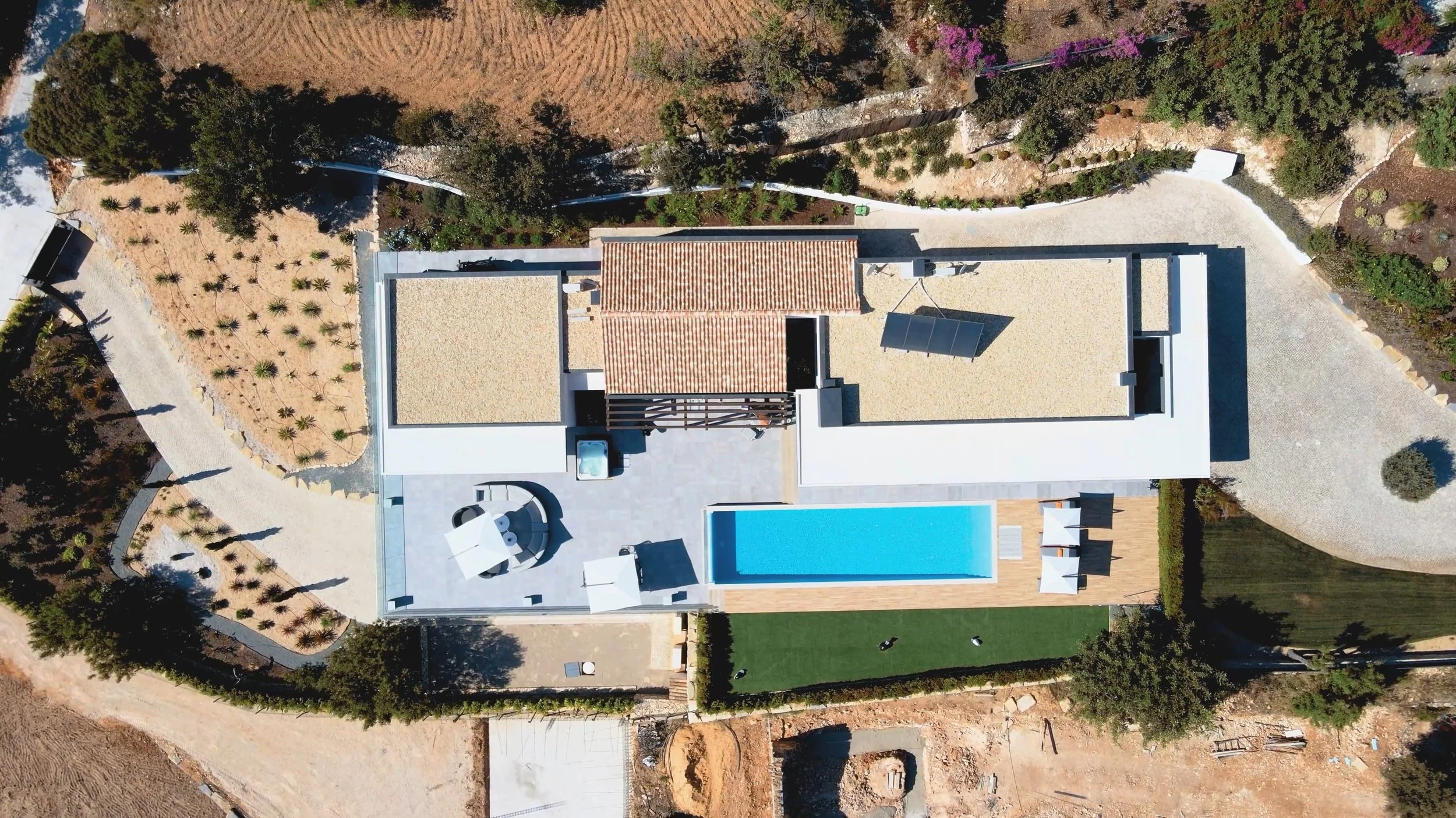 Vista aérea de uma residência moderna com piscina, área de lazer com mesas, cadeiras, sombra de guarda-sóis, jardim com grama e uma área de jardim com plantas ao redor.