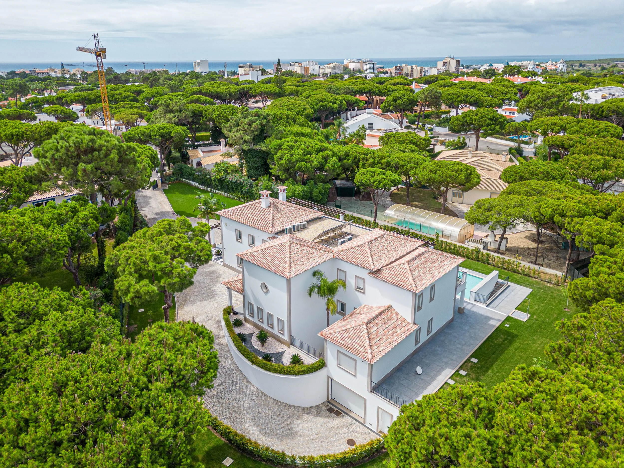 Vista aérea de uma casa moderna com telhado de telhas vermelhas, rodeada por árvores verdes, com piscina e jardim bem cuidado, em uma área residencial próxima ao mar.