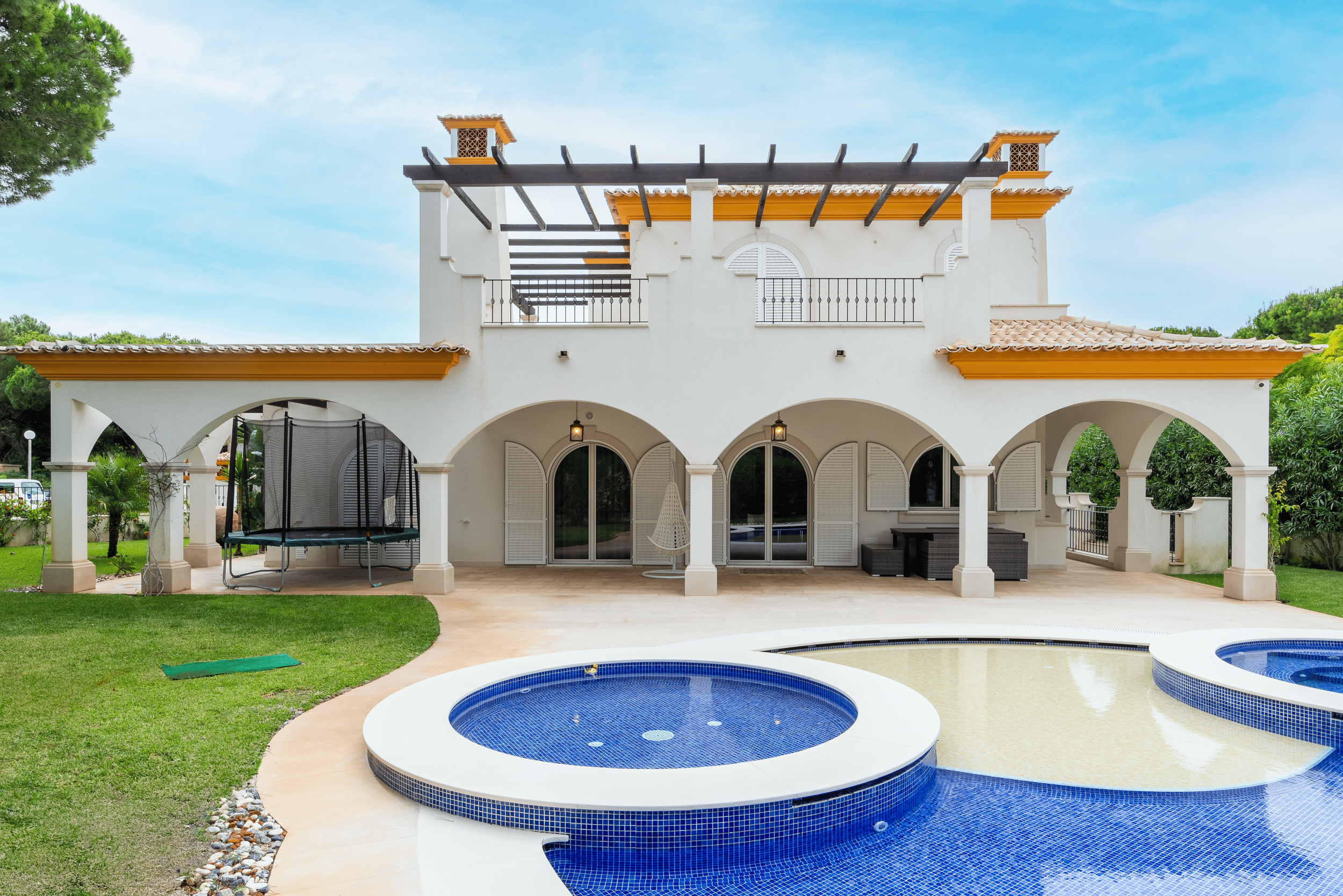 Casa grande com varanda com arco e piscina ao fundo, árvores e céu azul.