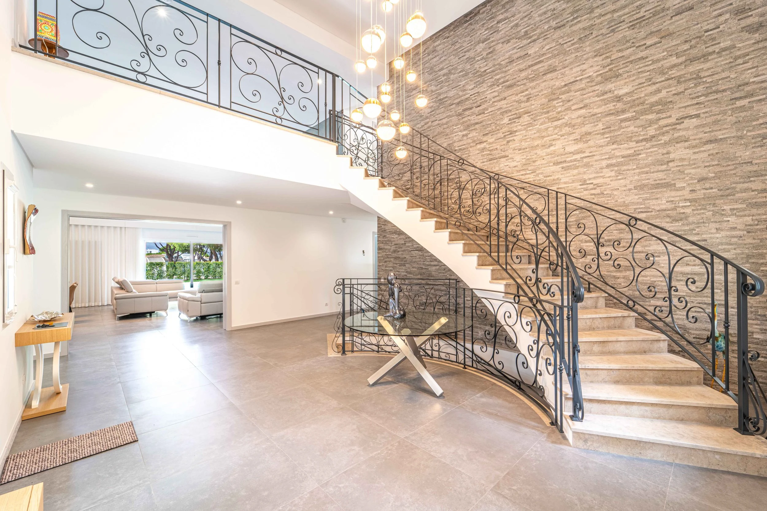 Sala de estar com escada de ferro detalhada, parede de pedra decorativa, iluminação moderna e espaço amplo com sofá branco vista de janela para o jardim.