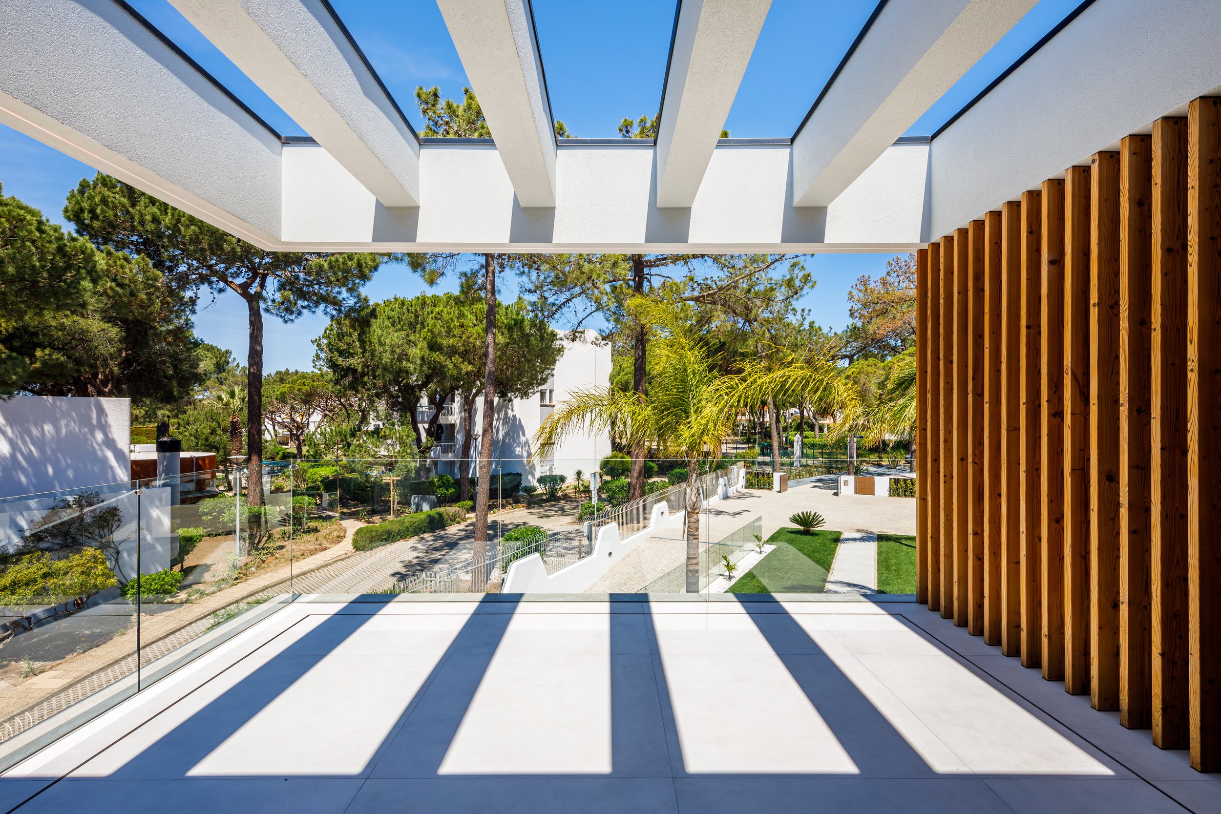Visão de varanda de um prédio com guarda-corpo de vidro, sombra de vigas no piso, árvores e jardim ao ar livre sob céu azul