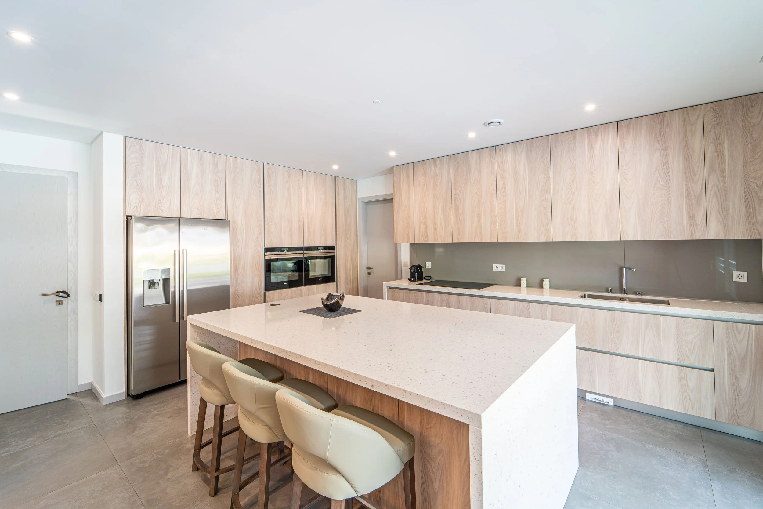 Cozinha moderna com armários de madeira clara, ilha central com superfície de pedra, cadeiras ao redor, geladeira inox, forno embutido e pia com torneira.