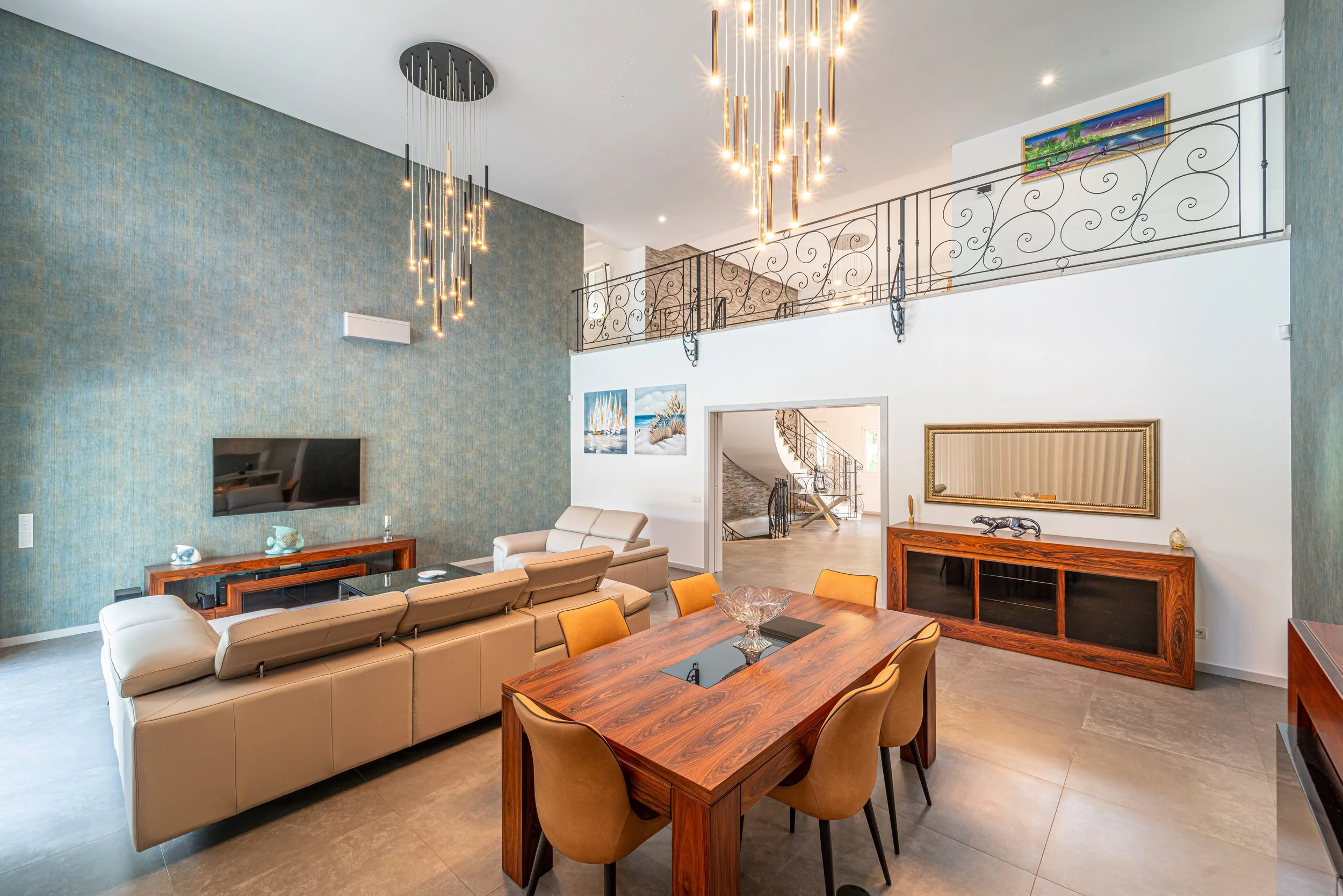 Sala de estar com painel azul na parede, sofá bege, mesa de jantar de madeira, cadeiras bege, aparador de madeira, televisão de tela plana na parede, iluminação moderna no teto, quadros na parede, escada de madeira e ferro, varanda com corrimão de ferro