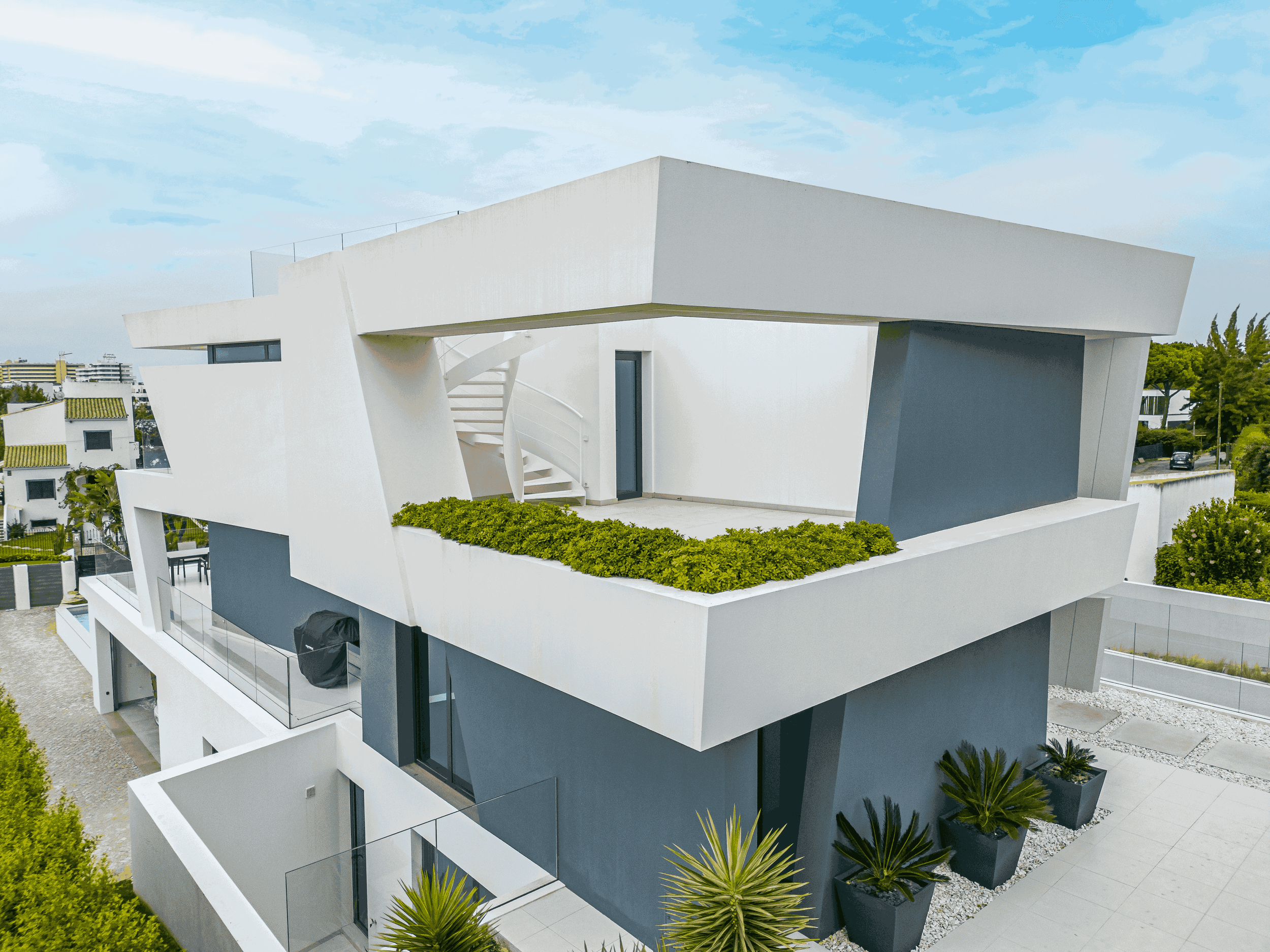 Edifício moderno de cores branco e cinza, com fachada angular e varanda com plantas verdes, em área urbana.