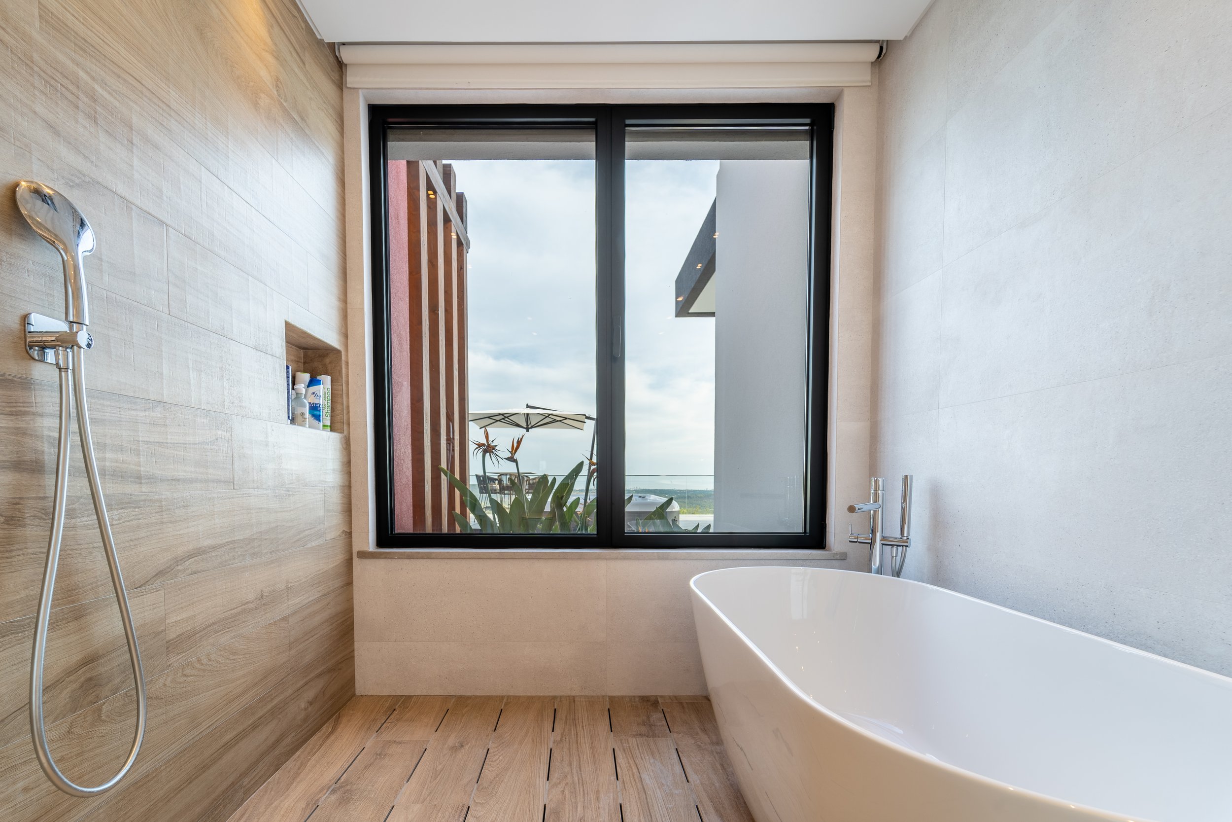 Banheira branca ao lado de uma janela grande em um banheiro com paredes de pedra e piso de madeira, com vista para um espaço externo com plantas, guarda-sol e céu parcialmente nublado.