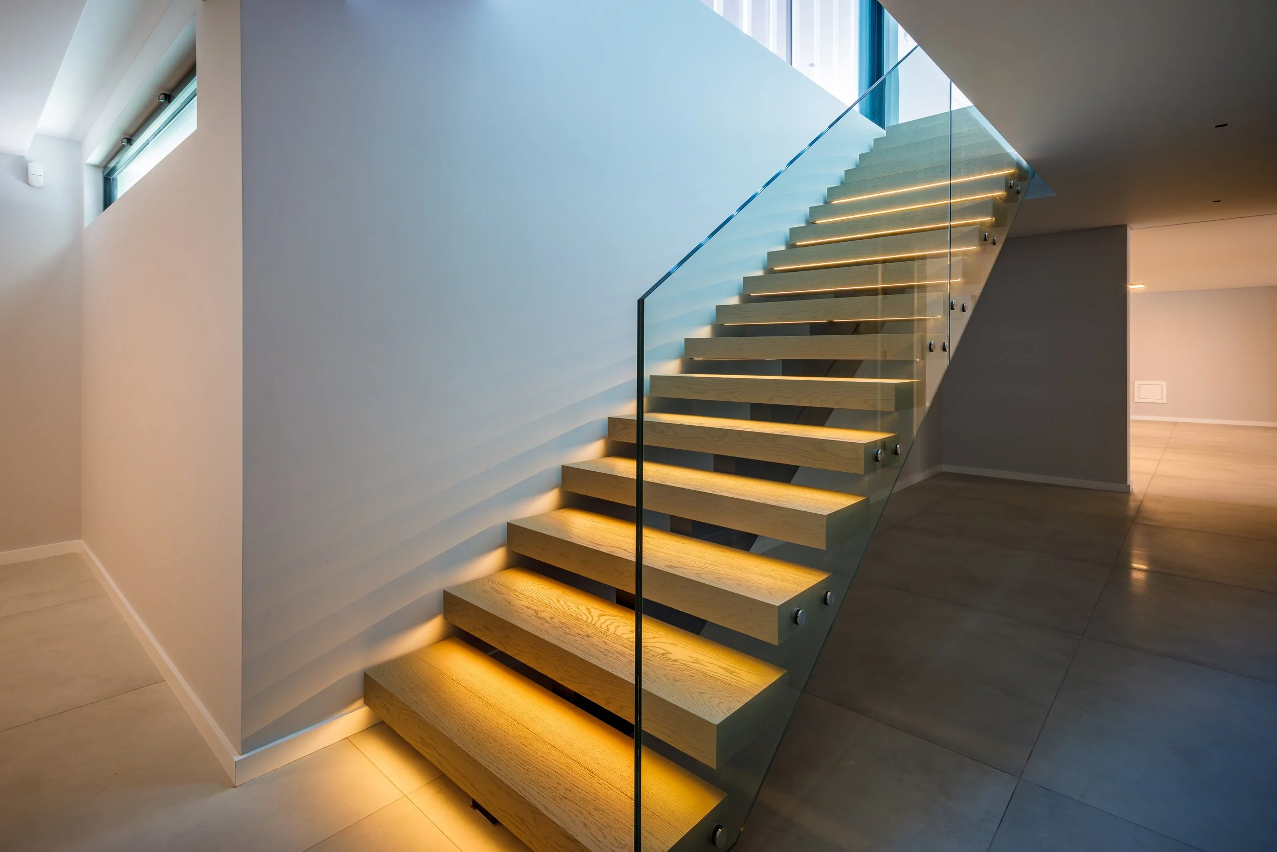 Escada moderna de madeira com iluminação embutida, com guarda-corpo de vidro, dentro de uma casa com paredes brancas e piso de azulejo.