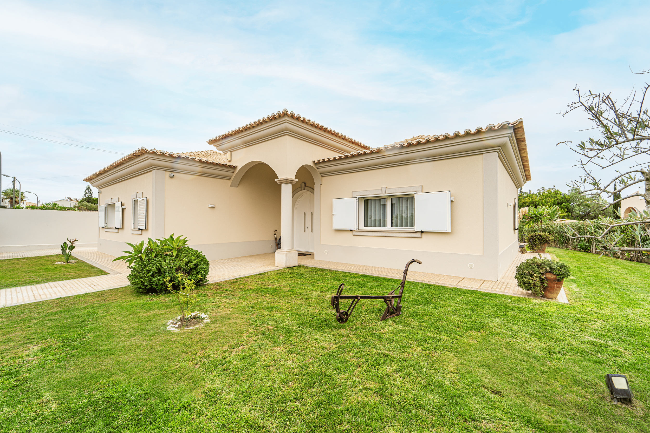 Casa de cor bege com telhado de telhas avermelhadas, janelas com persianas brancas, jardim com grama bem cuidado, plantas em vasos e um banco de madeira danificado no gramado.