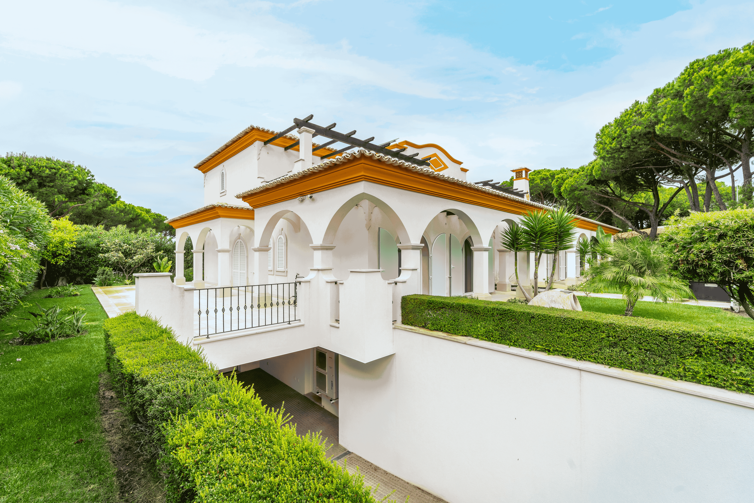 Casa branca com caixilharia de ferro preto, cercada por jardim com árvores e arbustos verdes, em dia claro com céu parcialmente nublado.