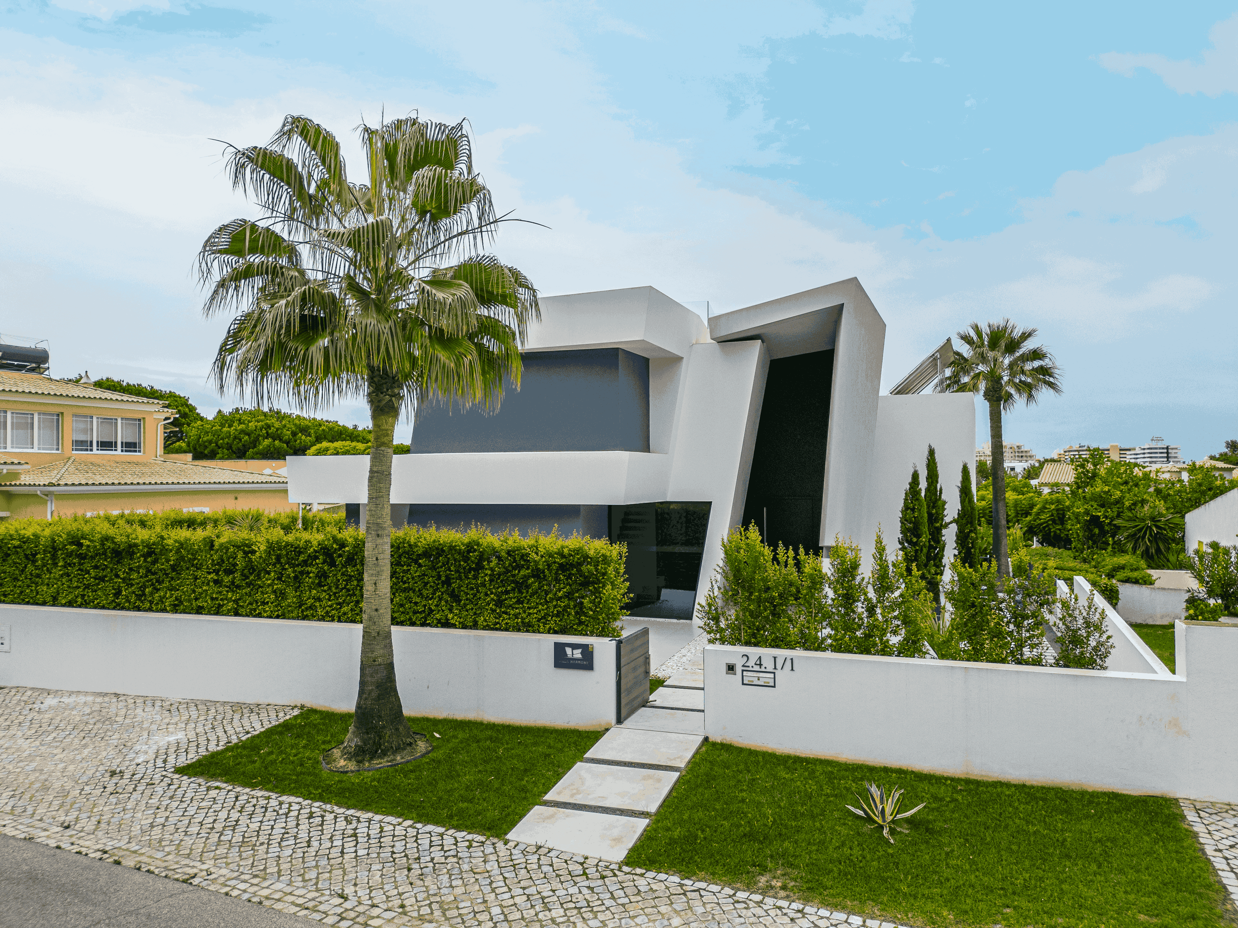 Casa moderna com arquitetura angular branca, cercada por um muro branco, com árvores de palmeira e arbustos verdes, sob céu parcialmente nublado.