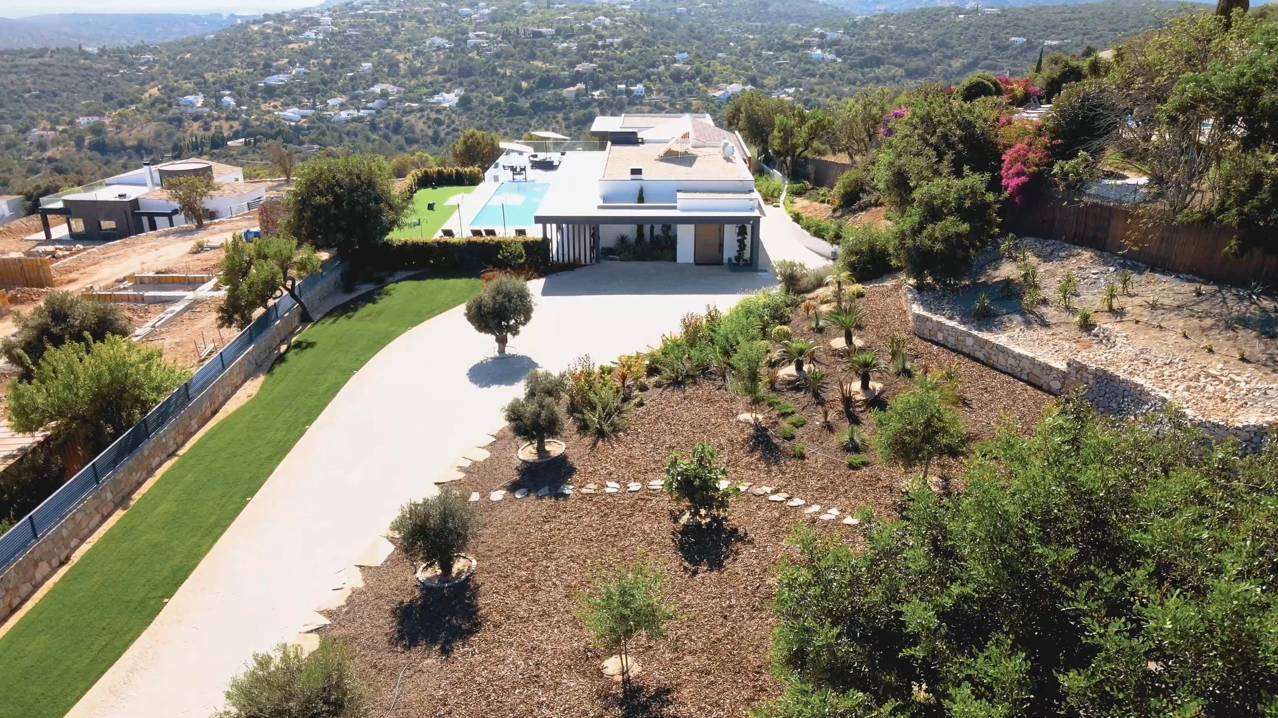 Imagem aérea de uma casa moderna com um jardim bem cuidado, piscina e árvores ao redor, em uma área com topo de morro.