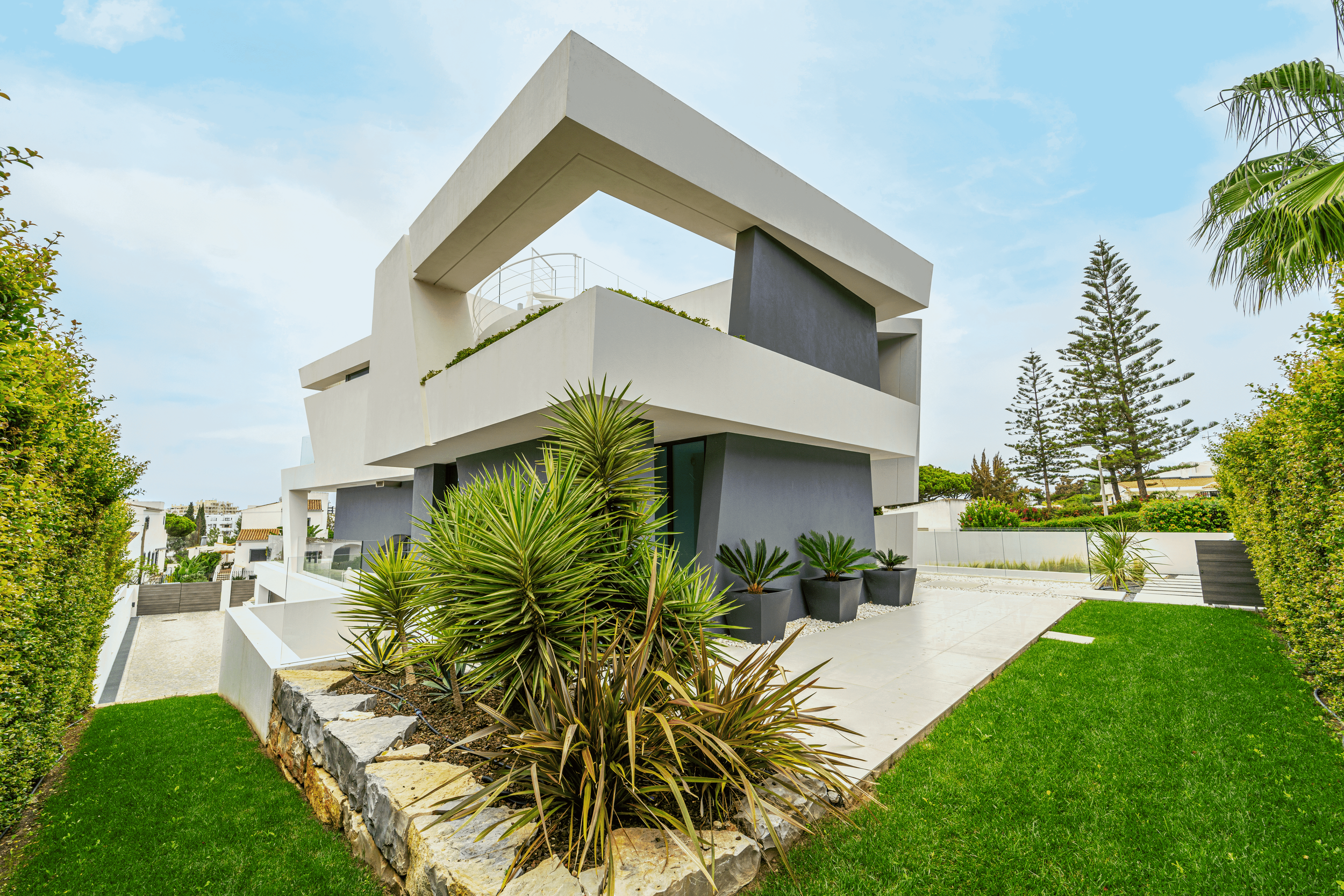 Imagem de uma casa moderna de arquitetura contemporânea, com elementos geométricos, paredes brancas e detalhes escuros, rodeada por jardim com plantas suculentas e gramado bem cuidado, sob céu claro com nuvens leves.