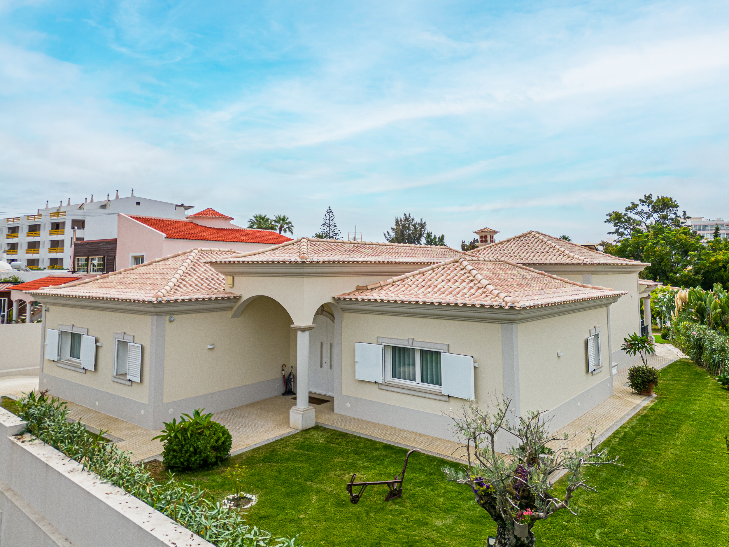Casa com telhado de telhas cerâmicas, paredes claras, janelas com persianas brancas, jardim bem cuidado com árvores e grama verde, e céu com nuvens.