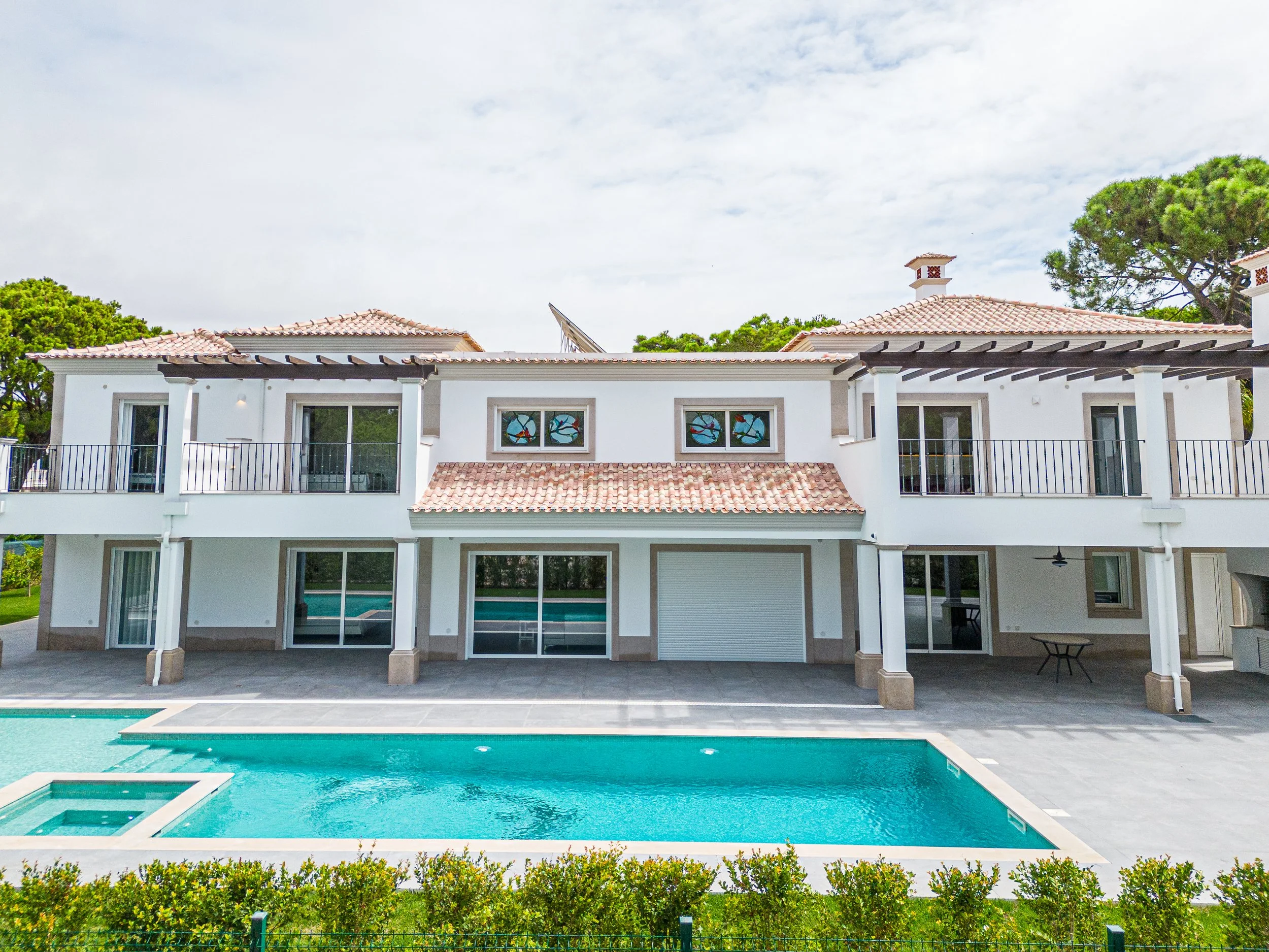 Casa moderna com teto de telhas, varanda com colunas, várias janelas com vitrais, piscina ao fundo e árvores ao redor.