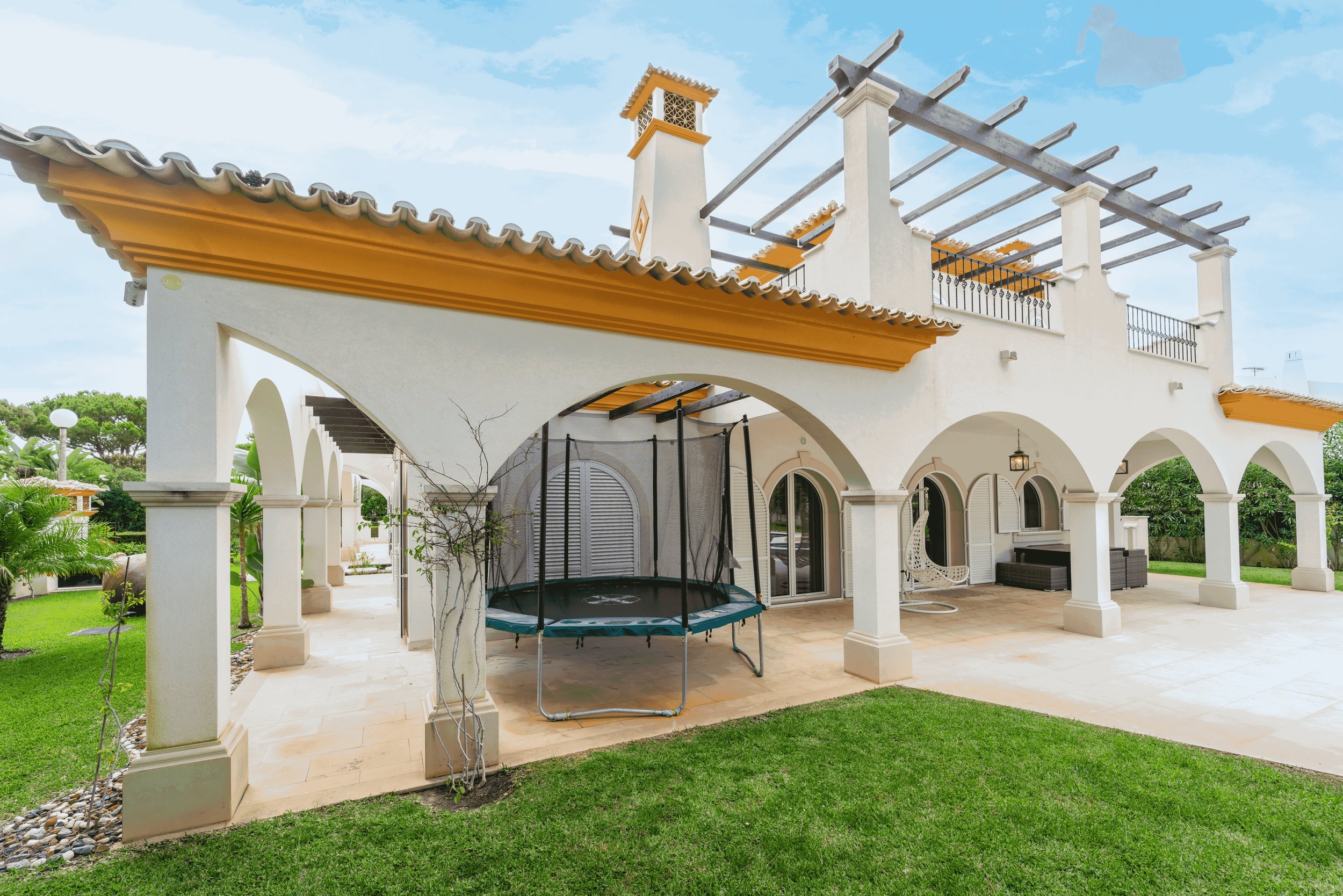 Varanda de casa com arcos, trampolim, cadeira de balanço, sofá de canto, jardim bem cuidado com gramado e árvores ao redor.