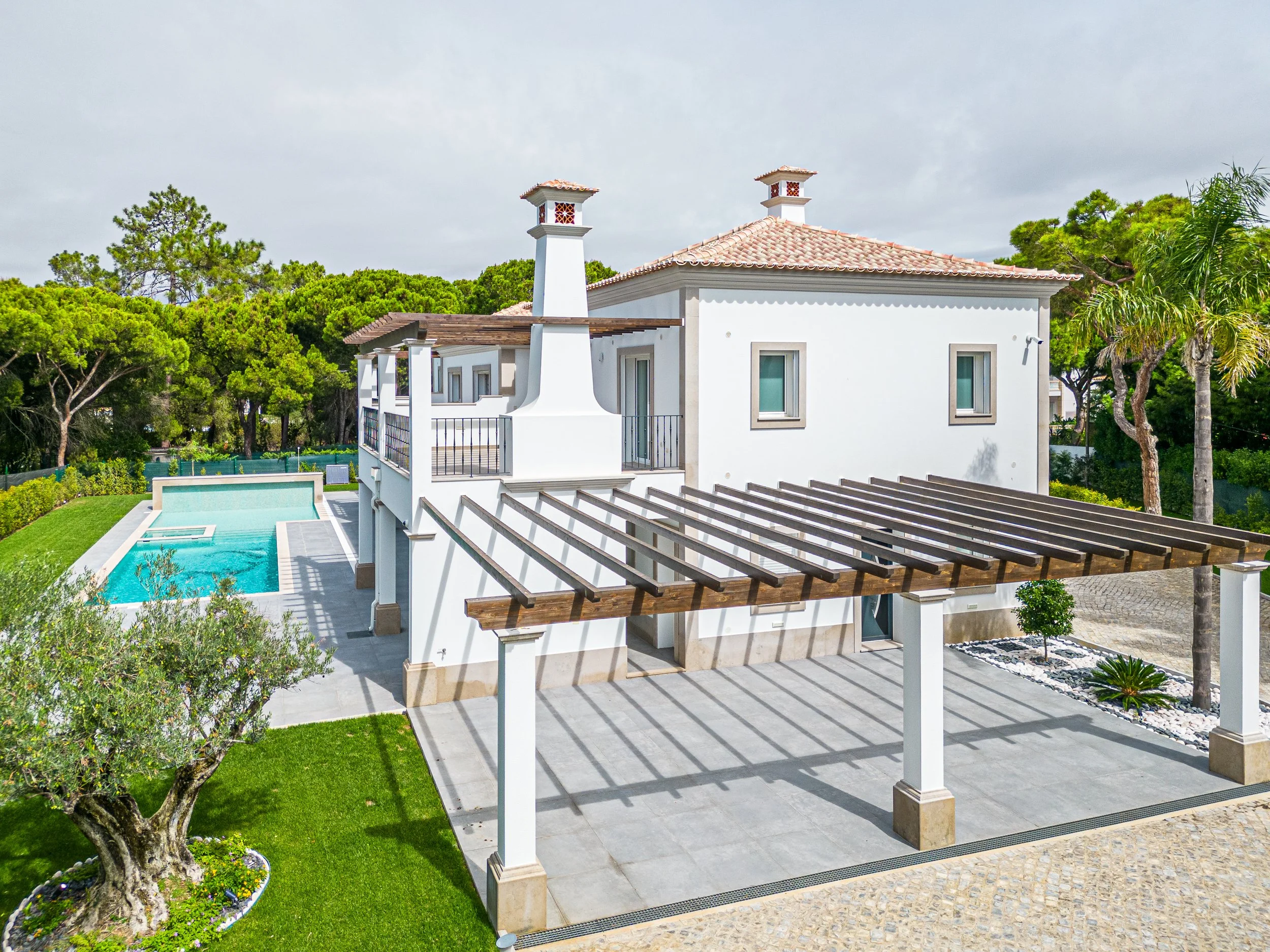 Casa branca com telhado de barro vermelho, piscina ao fundo, jardim bem cuidado e estrutura de madeira em pátio externo.