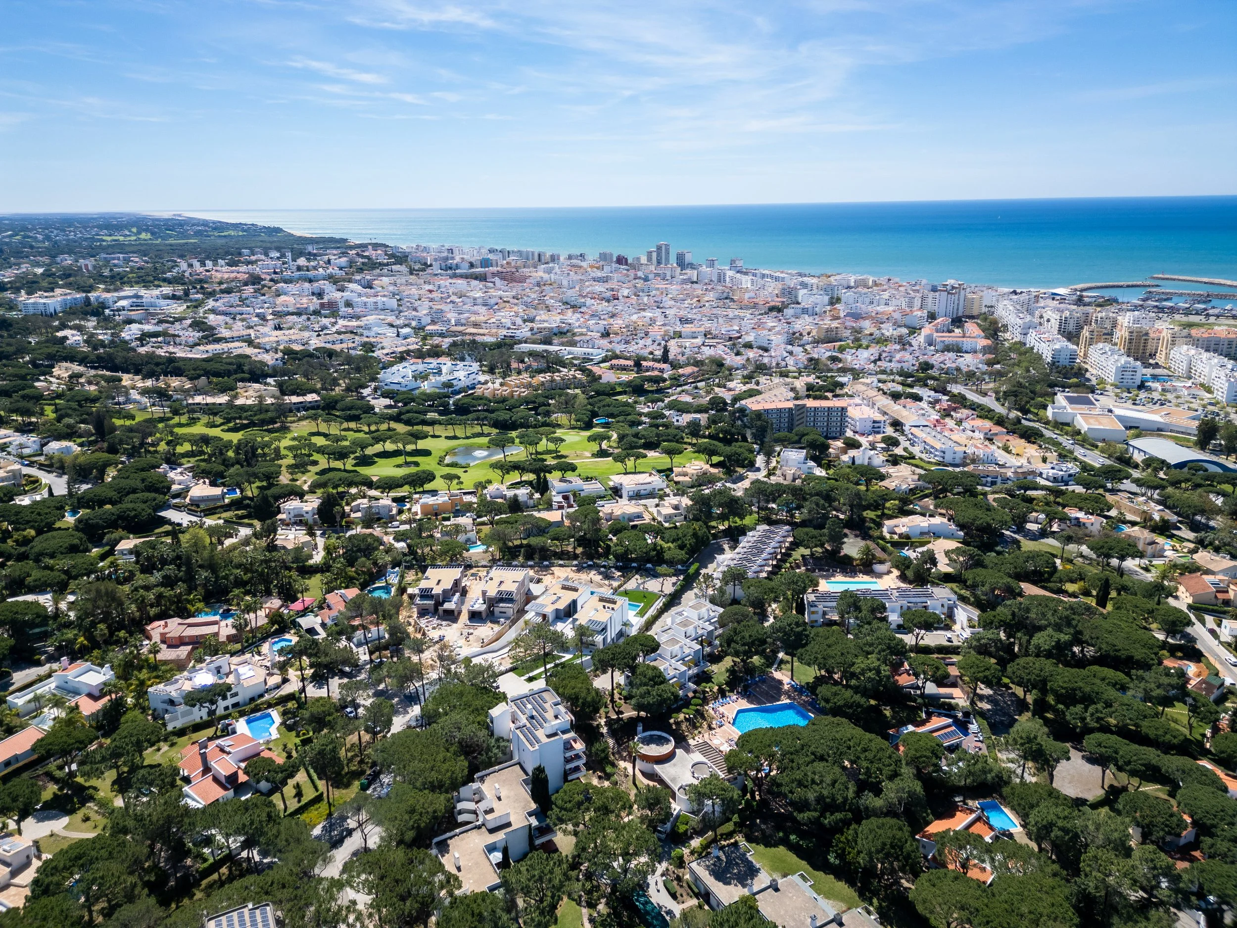 Vista aérea de uma cidade costeira com prédios, áreas verdes, piscinas e o mar ao fundo.
