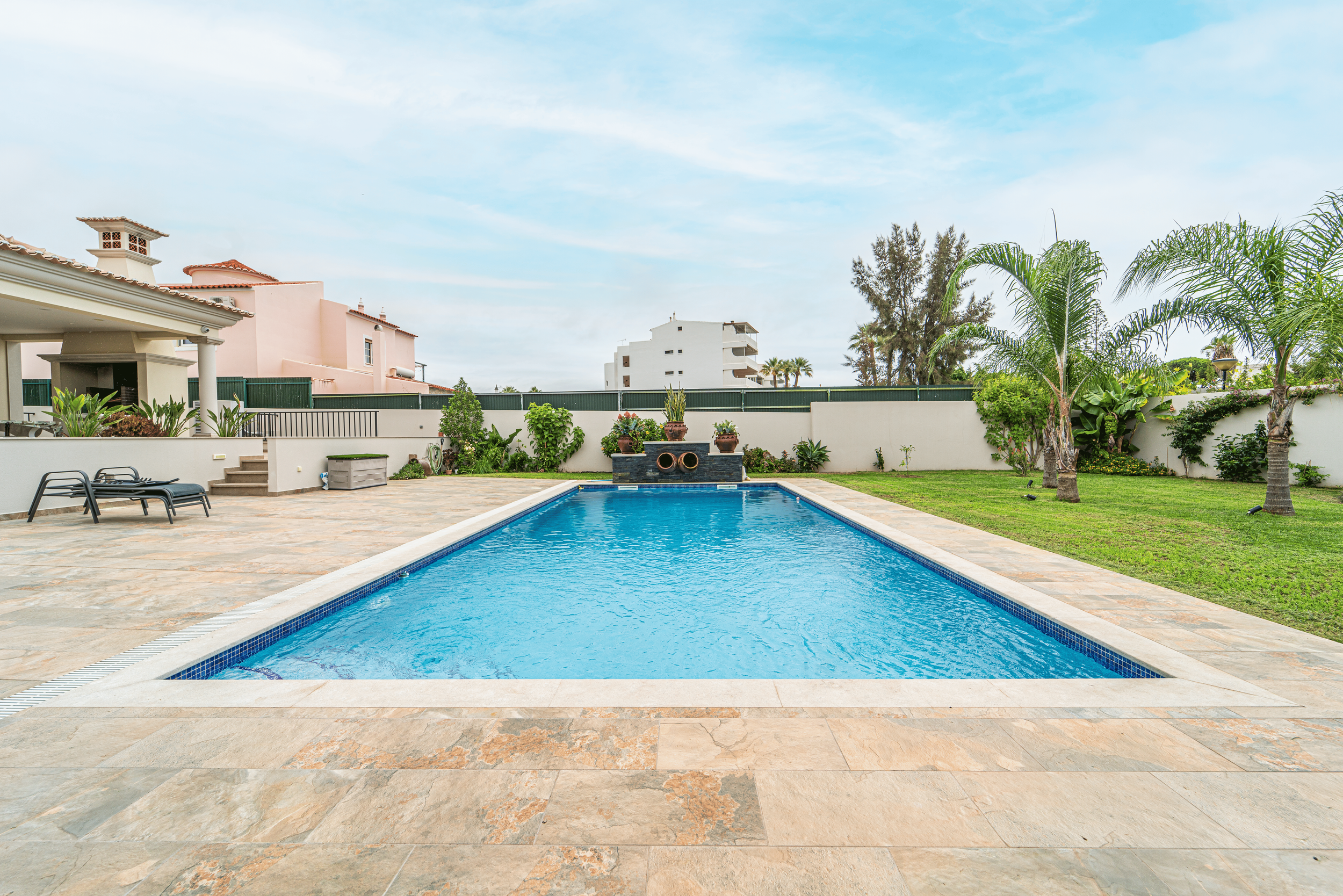 Piscina rectangular com borda de pedra, espreguiçadeiras ao lado, área de jardim com gramado, árvores de palmeira e plantas, muro de fundo, céu azul com algumas nuvens.