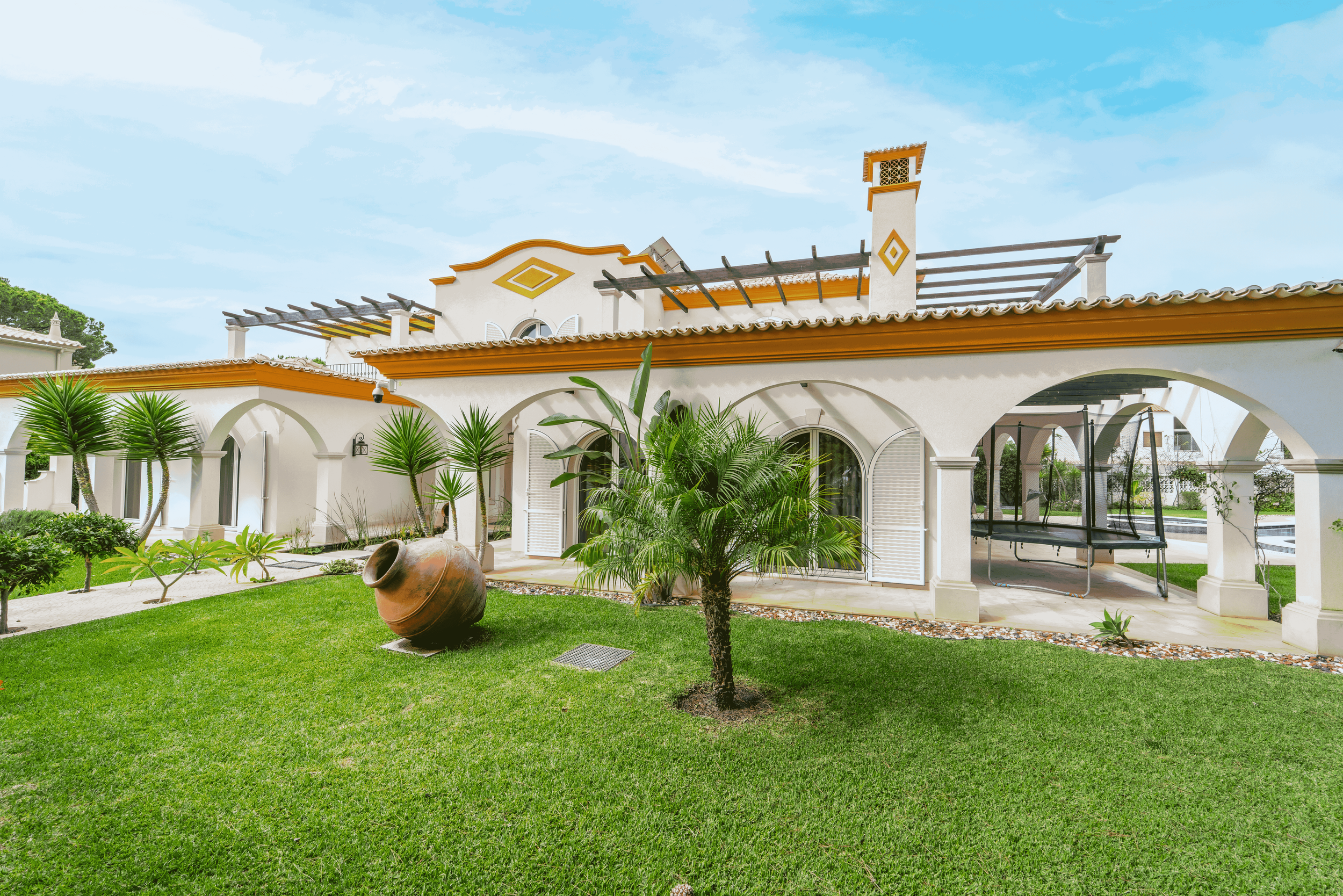Casa de estilo mediterrâneo com jardim bem cuidado, árvores tropicais e espaço ao ar livre com uma piscina ao fundo.