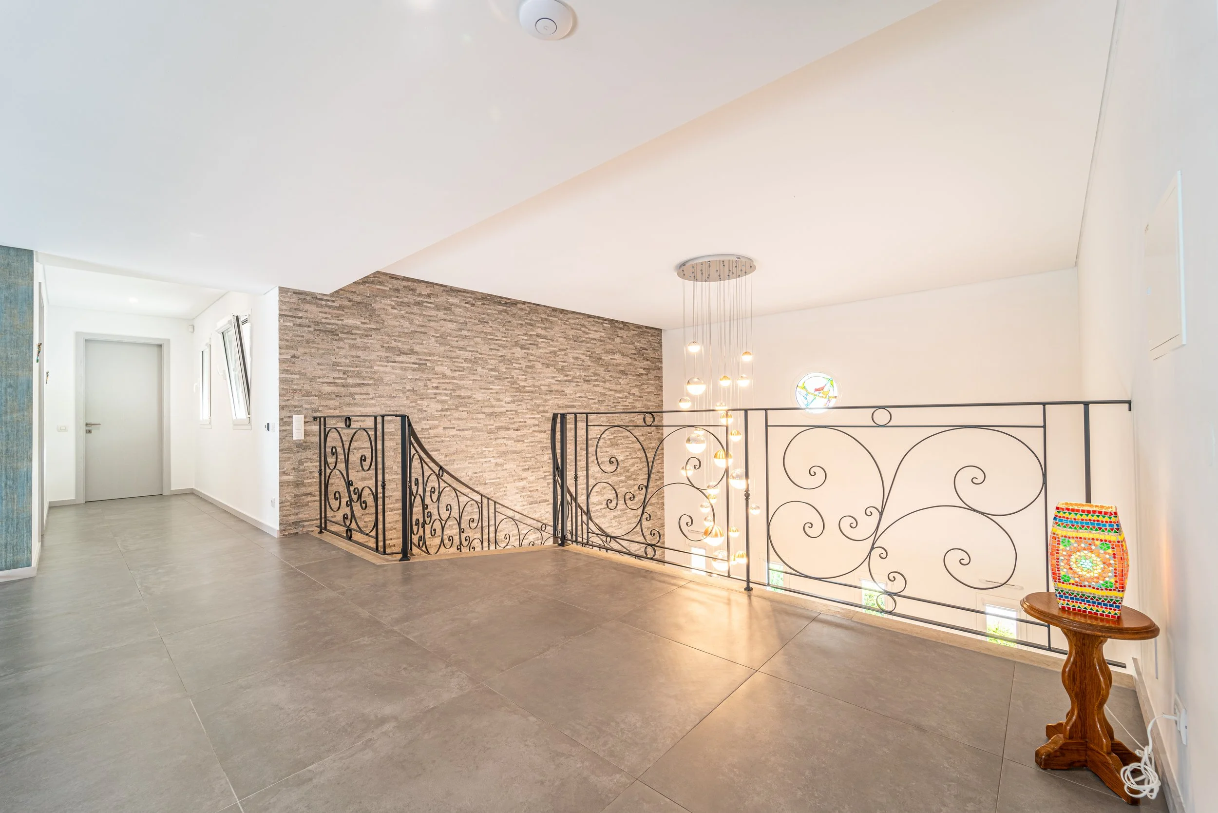 Vista de um espaço interno com piso de cerâmica cinza, parede de pedra, escada com corrimão de ferro decorativo e lustre de luz pendurado. Há uma mesa pequena com uma luminária colorida no canto à direita.
