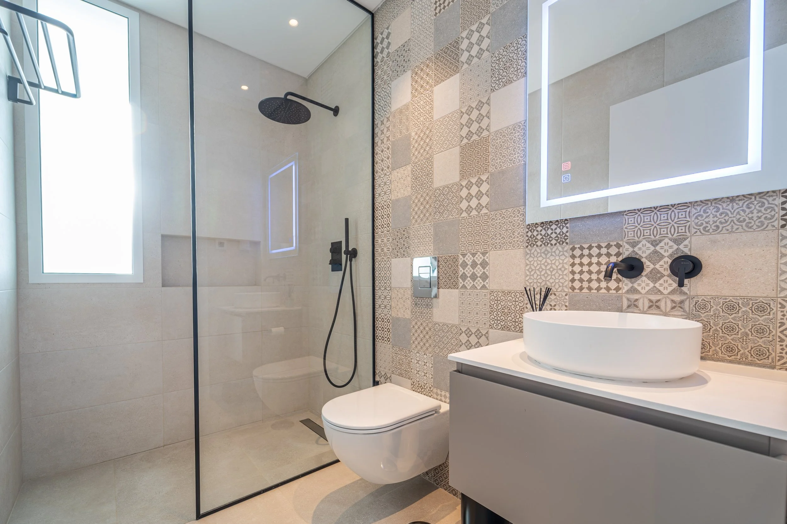 Banheiro moderno com parede de azulejos decorativos, pia de vaso sanitário branco, espelho grande com iluminação e área de chuveiro com vidro e ducha preta.