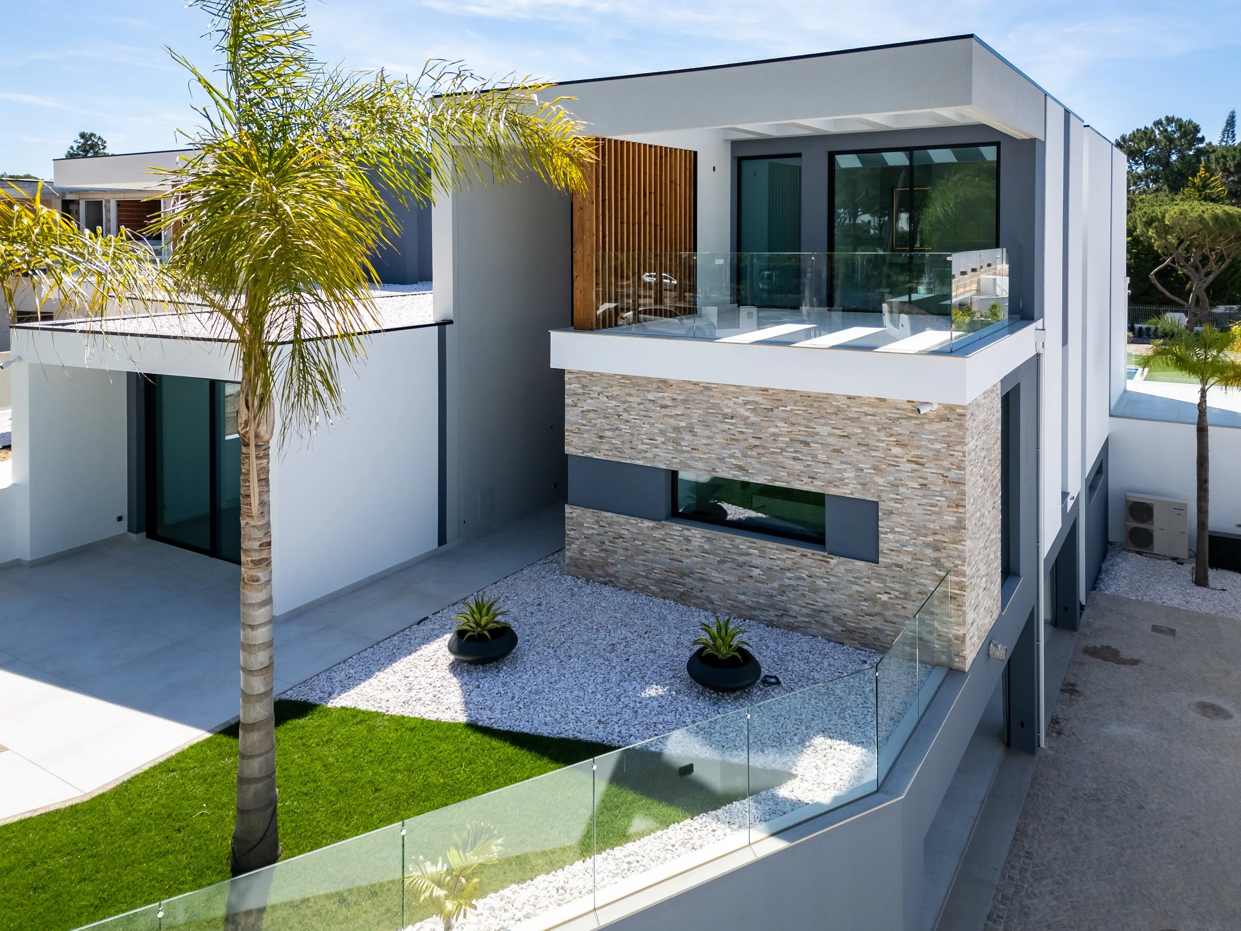Visual de uma casa moderna com fachada de revestimento de pedras, varanda com vidro e espaço externo com gramado, plantas decorativas e parede branca.