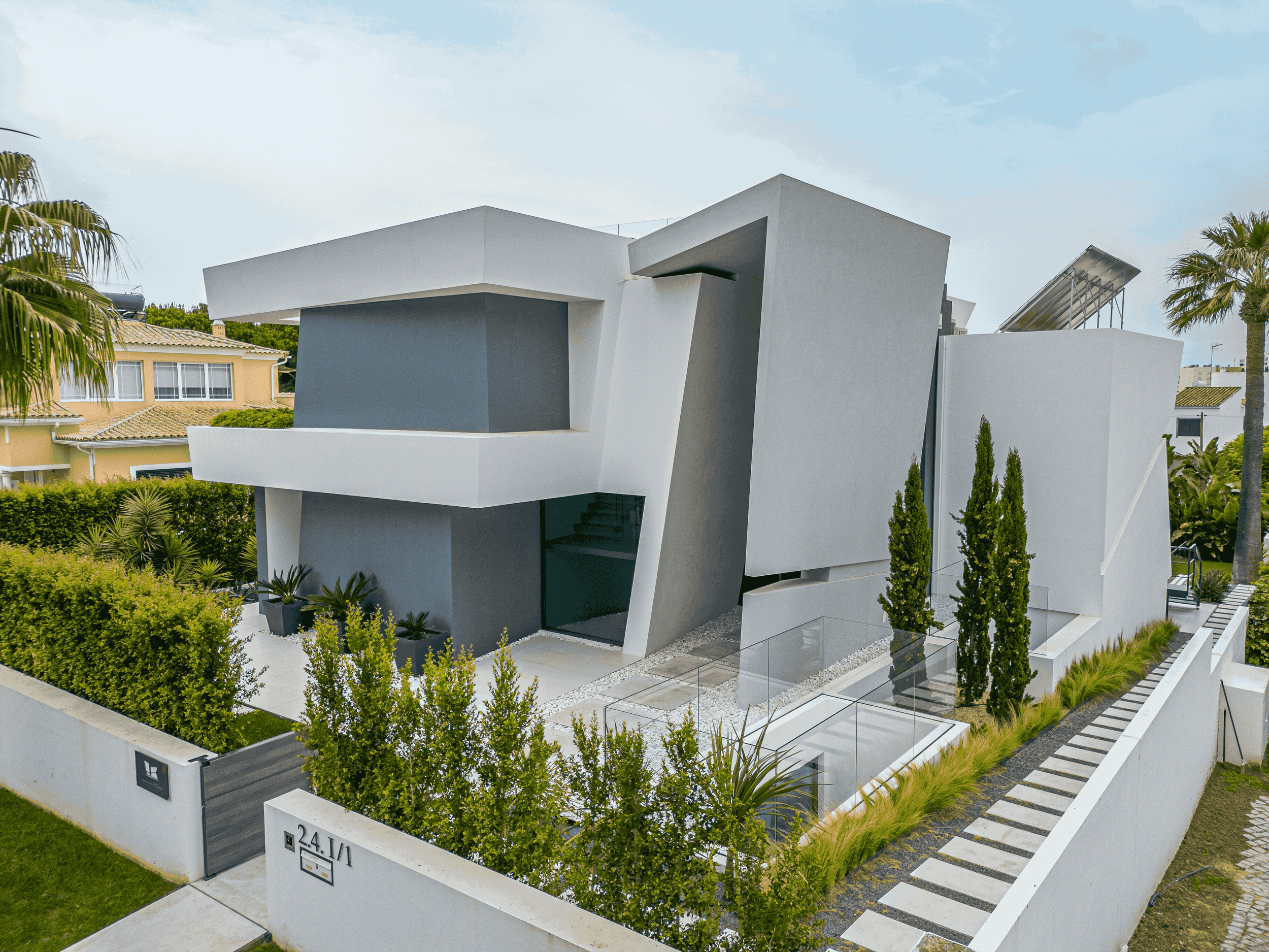 Prédio moderno de arquitetura contemporânea com linhas geométricas, paredes brancas e cinza, janelas amplas de vidro, jardim bem cuidado com plantas altas e árvores, e caminho pavimentado ao lado.
