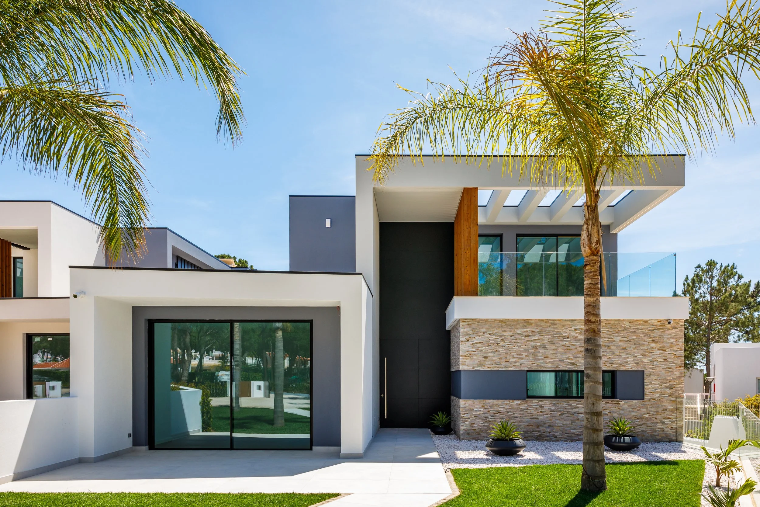Imagem de uma casa moderna com fachada de pedras, paredes brancas e janelas grandes. Há árvores e plantas ao redor, incluindo duas palmeiras, em um dia ensolarado com céu azul.