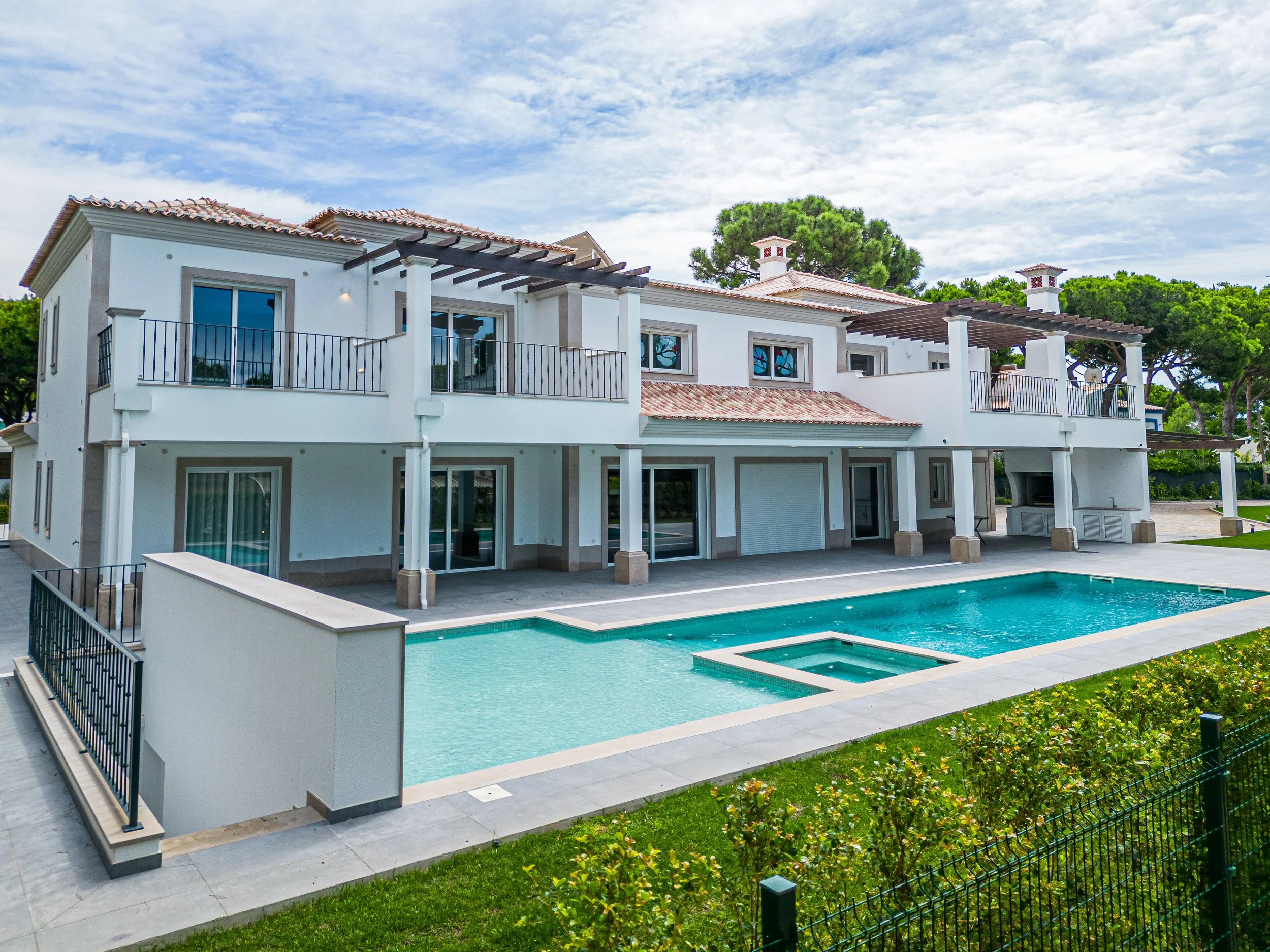 Residência moderna de duas andares com pátio e piscina ao ar livre, cercada por grama e arbustos, sob céu parcialmente nublado.