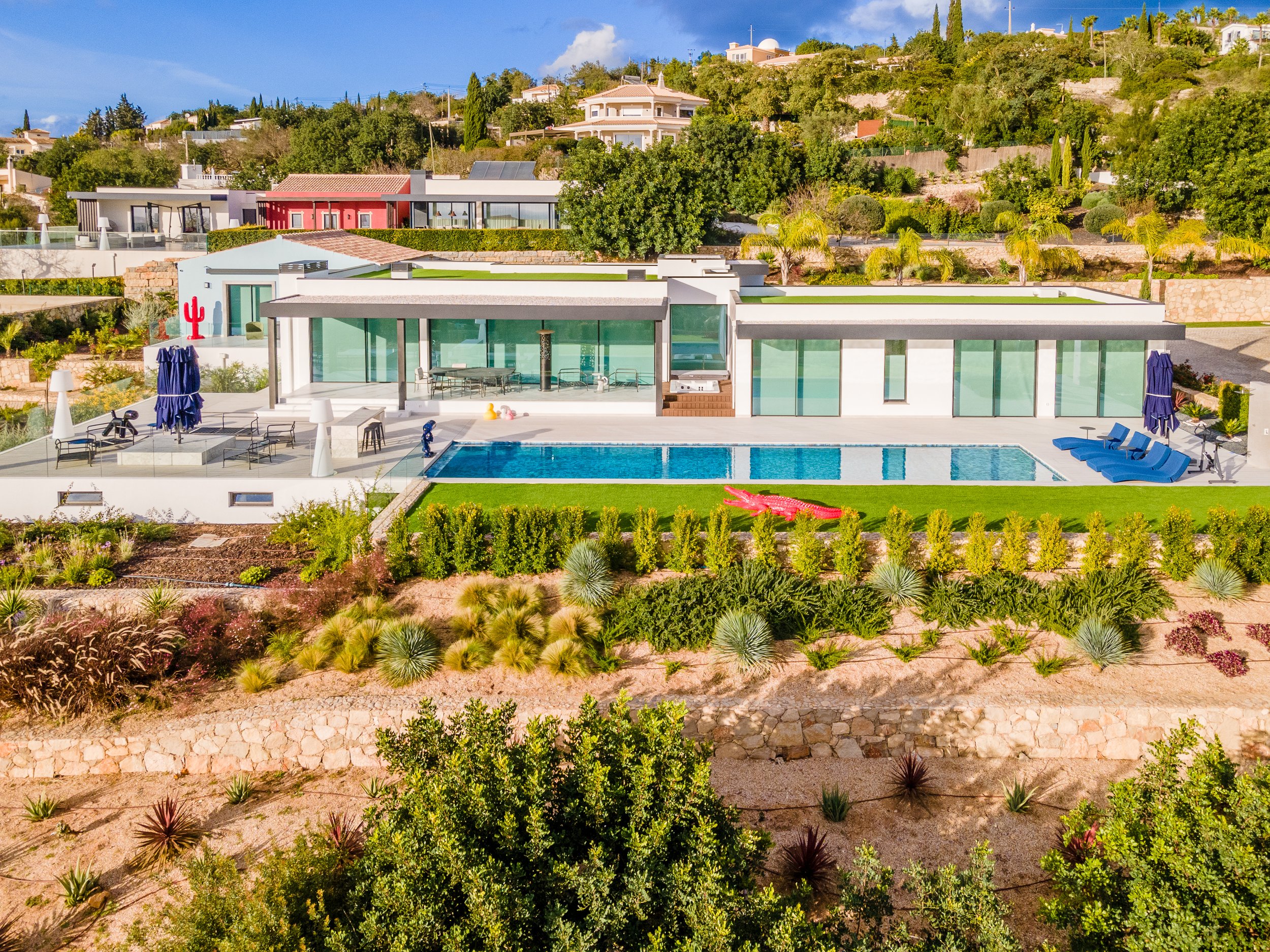 Casa moderna com piscina na área externa, espreguiçadeiras azuis e jardim bem cuidado com plantas de diferentes tamanhos e cores, em ambiente ensolarado.