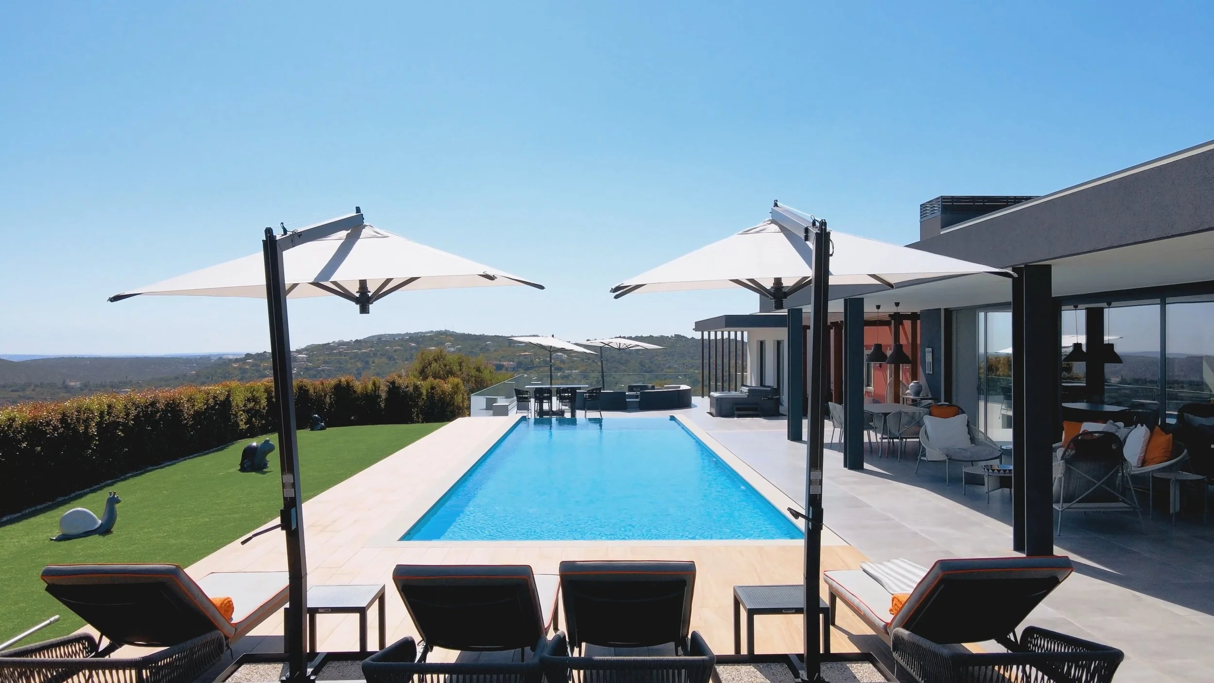 Piscina infinita com espreguiçadeiras, guarda-sóis brancos e área de lazer moderna ao redor, com vista para montanhas ao fundo.