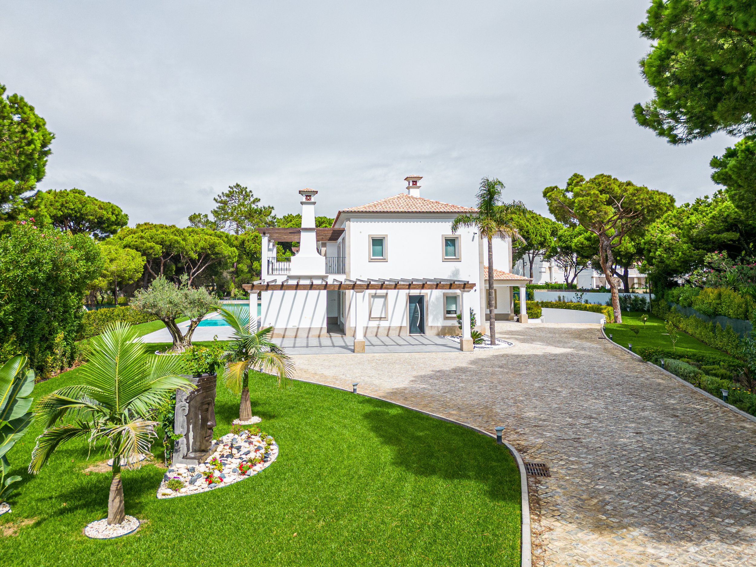 Casa branca com jardim bem cuidado e árvores ao redor, estrada de pedra na frente.