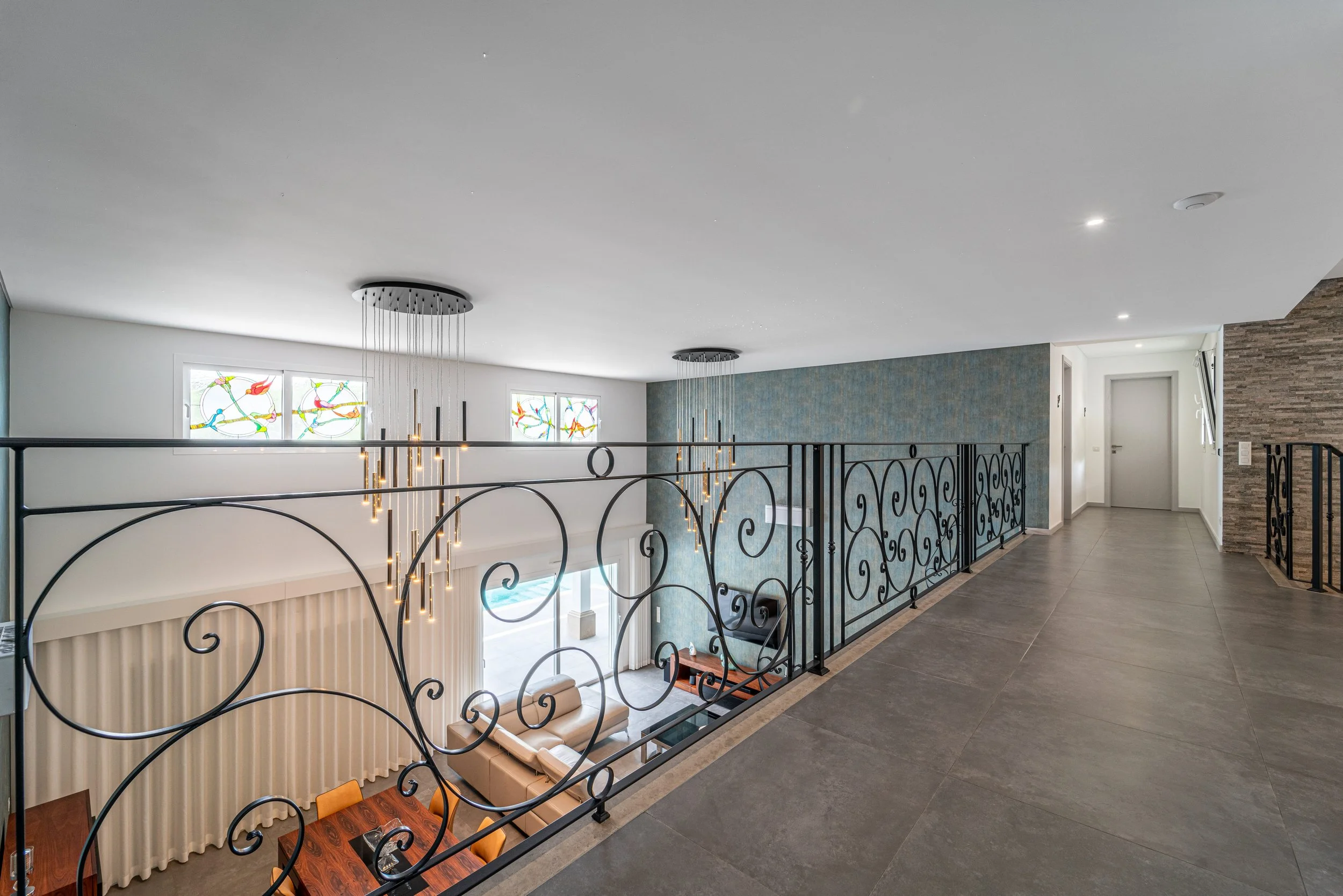 Corredor superior de uma casa moderna com corrimão de ferro trabalhado, luminária pendente de vários fios com luzes pequenas, janelas com vitrais coloridos, parede azul com textura e piso de cerâmica cinza. Vislumbre de uma sala com móvel de couro bege e mesa de jantar de madeira.