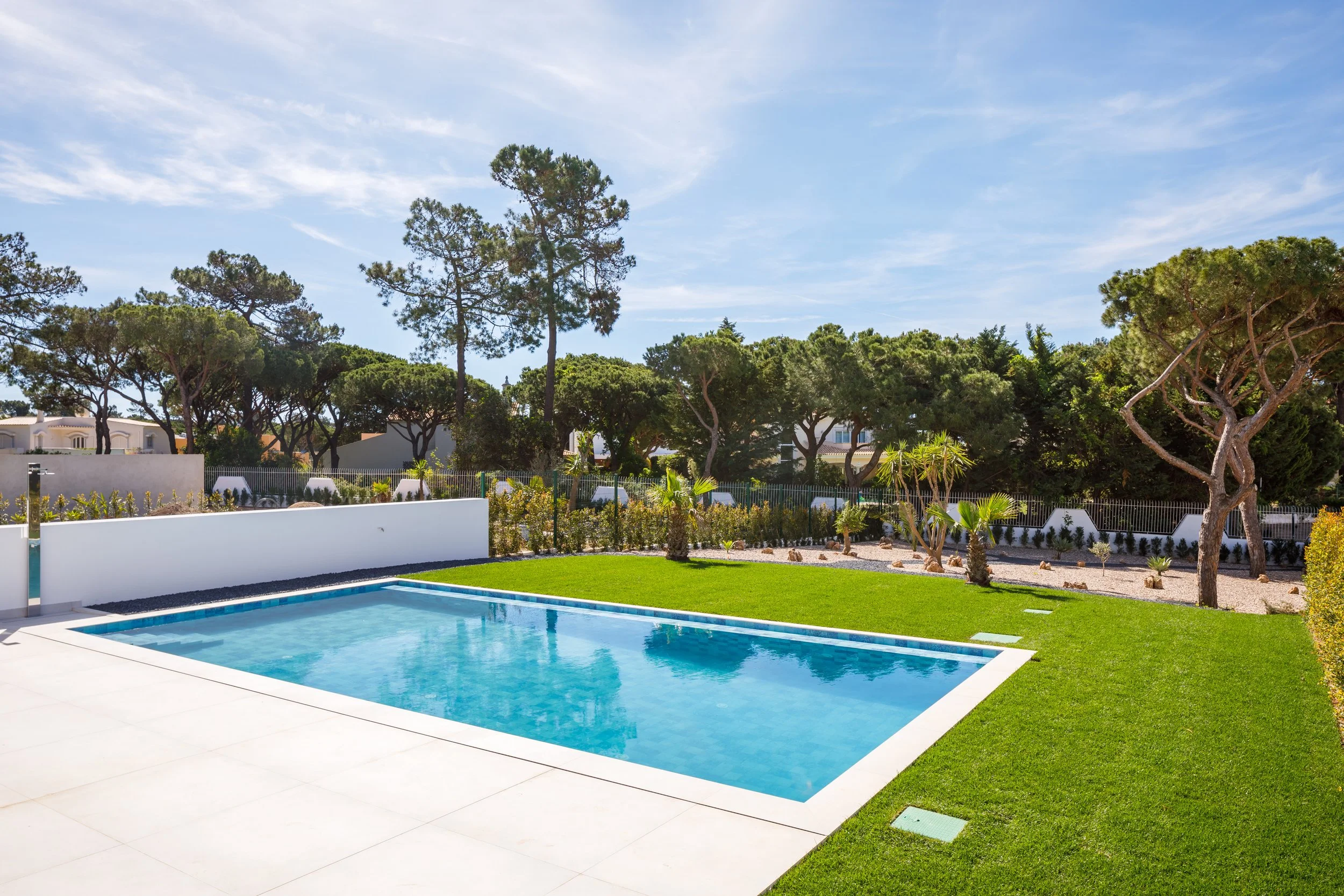Piscina de água azul clara em um jardín com grama verde, árvores ao fundo, céu ensolarado e árvores de grande porte ao longe.
